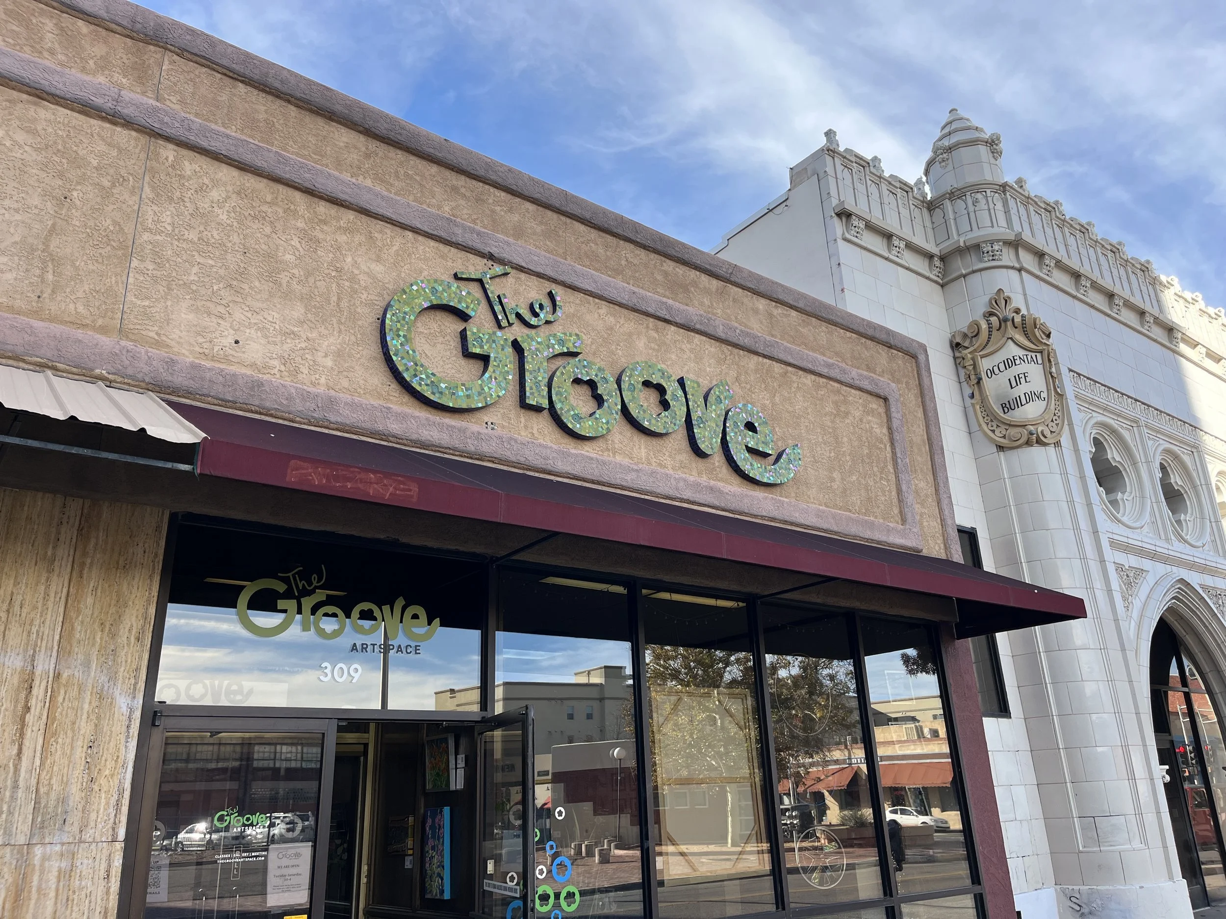 The exterior of an art space called The Grove, located at 309, with a sign on the building, glass doors, and a maroon awning. The building next door is the Occidental Life Building with ornate architecture and a sign.