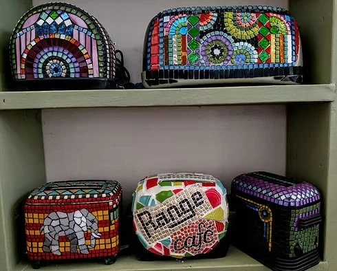 Six colorful mosaic-style bags and containers displayed on a white shelf, including two bags on the top shelf and four containers or boxes on the bottom shelf.