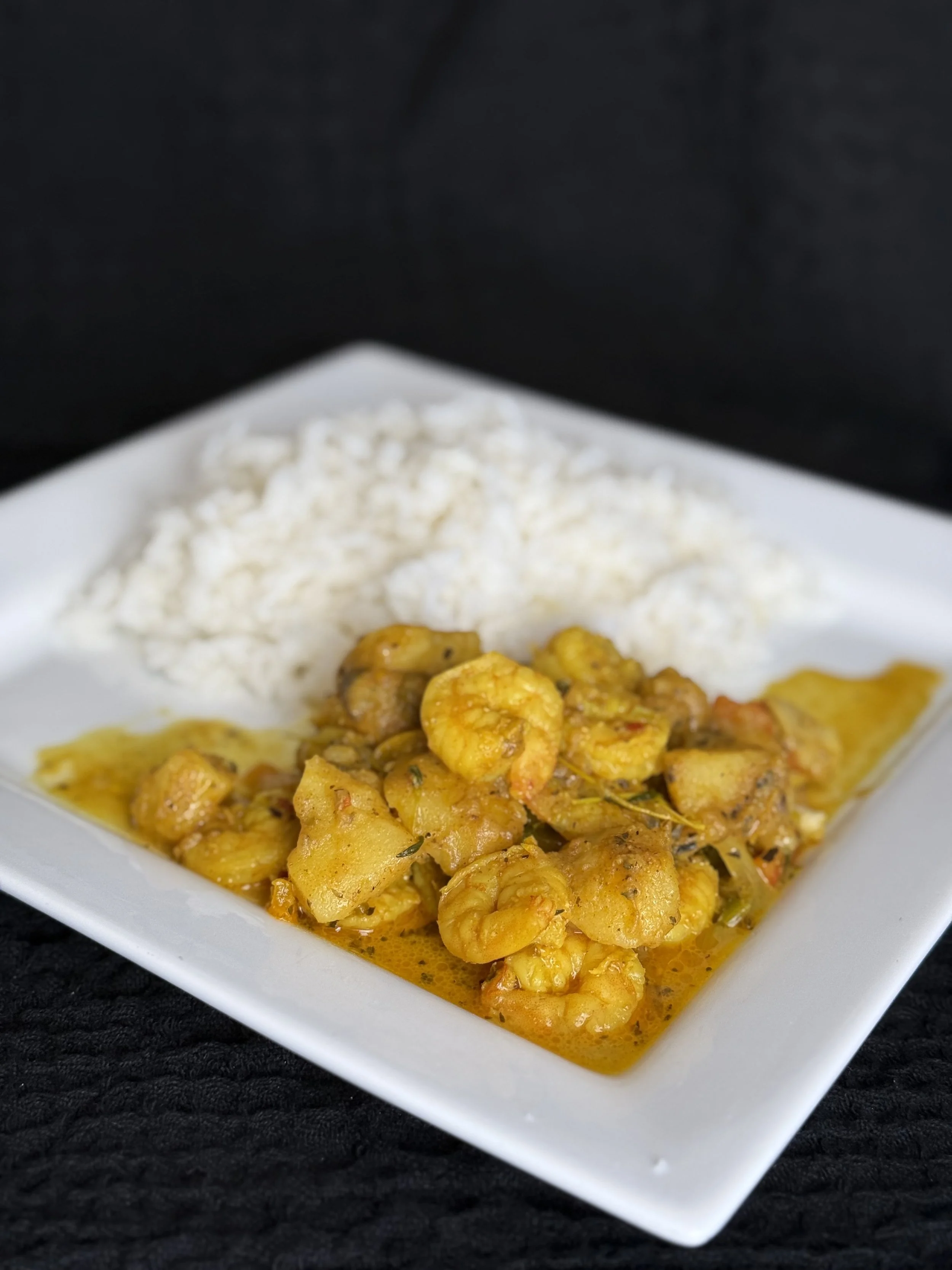 A square white plate with cooked shrimp in yellow curry sauce and a mound of white rice against a dark background.