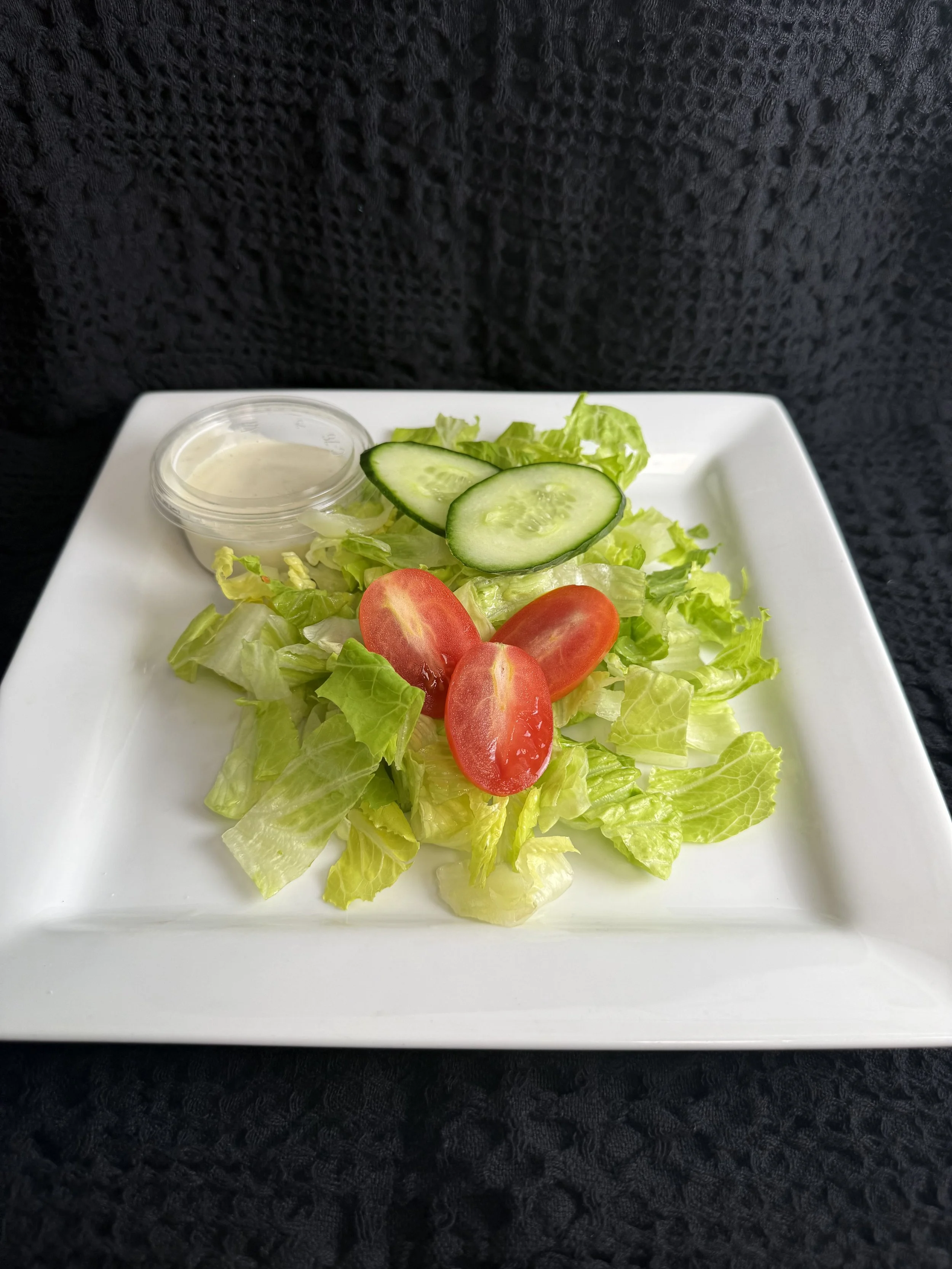 Fresh garden salad with lettuce, cherry tomatoes, cucumber slices, a small container of ranch dressing, and a side of creamy salad dressing on a white square plate, placed on a black textured surface.