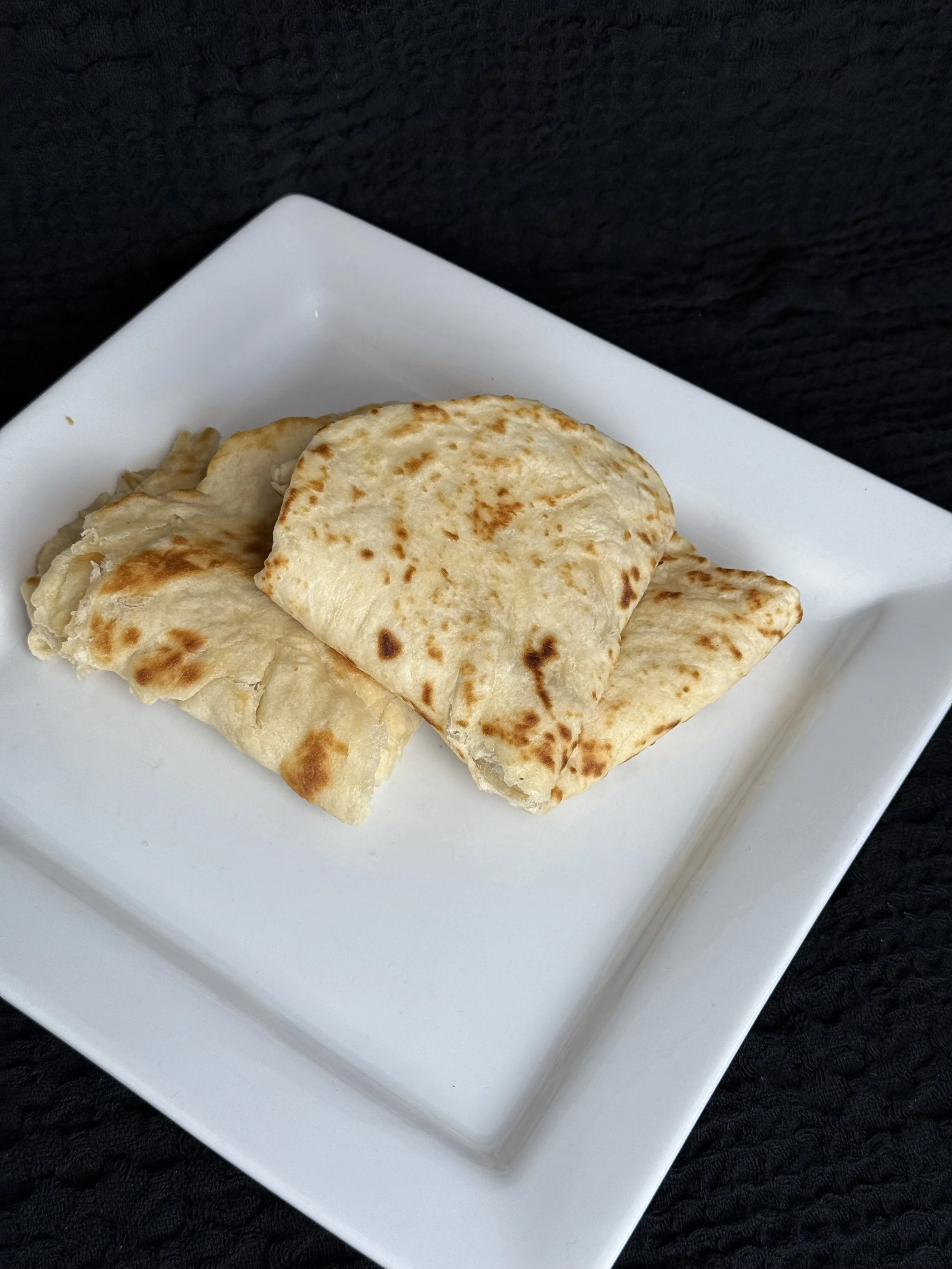 Two pieces of flatbread on a white square plate against a black textured background.