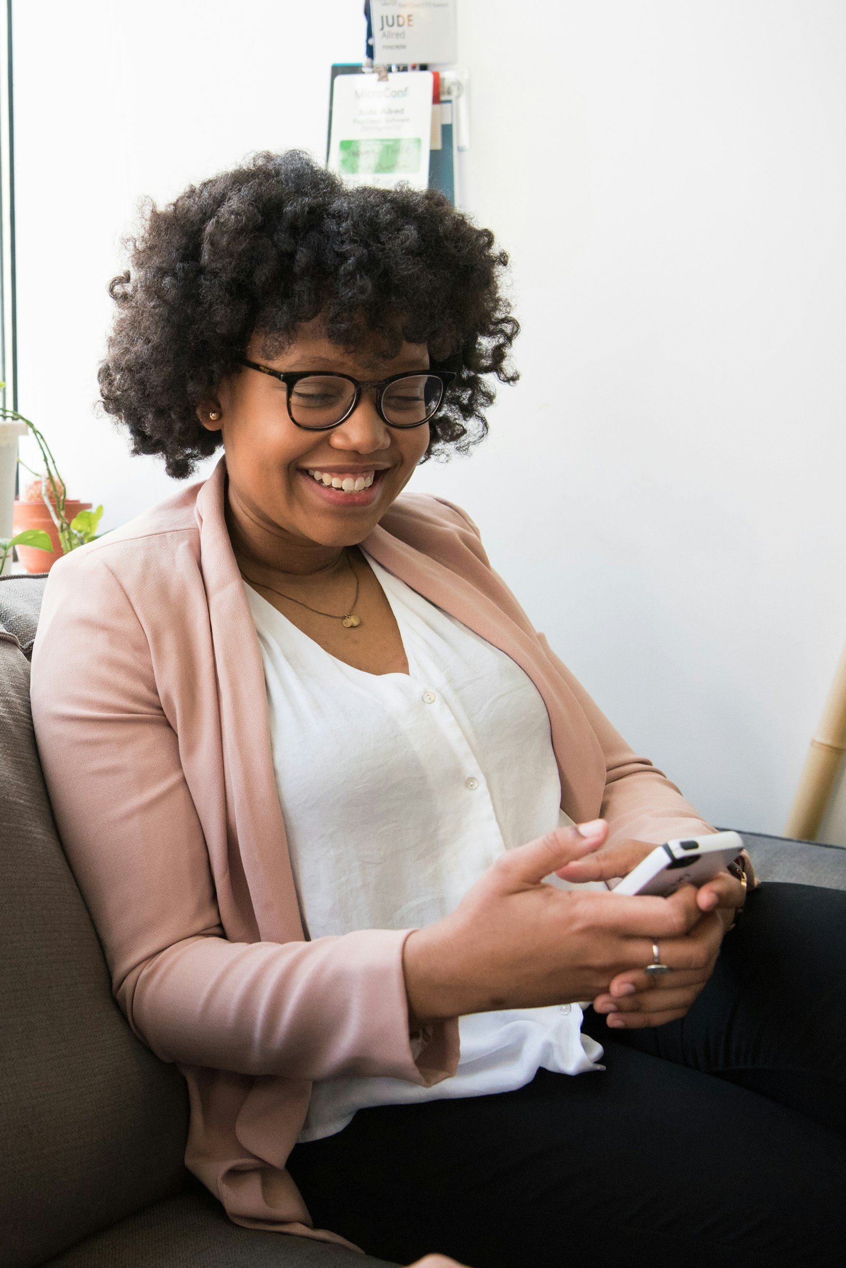 A woman in a white shirt and a pink sweater is smiling at her phone in her hand.