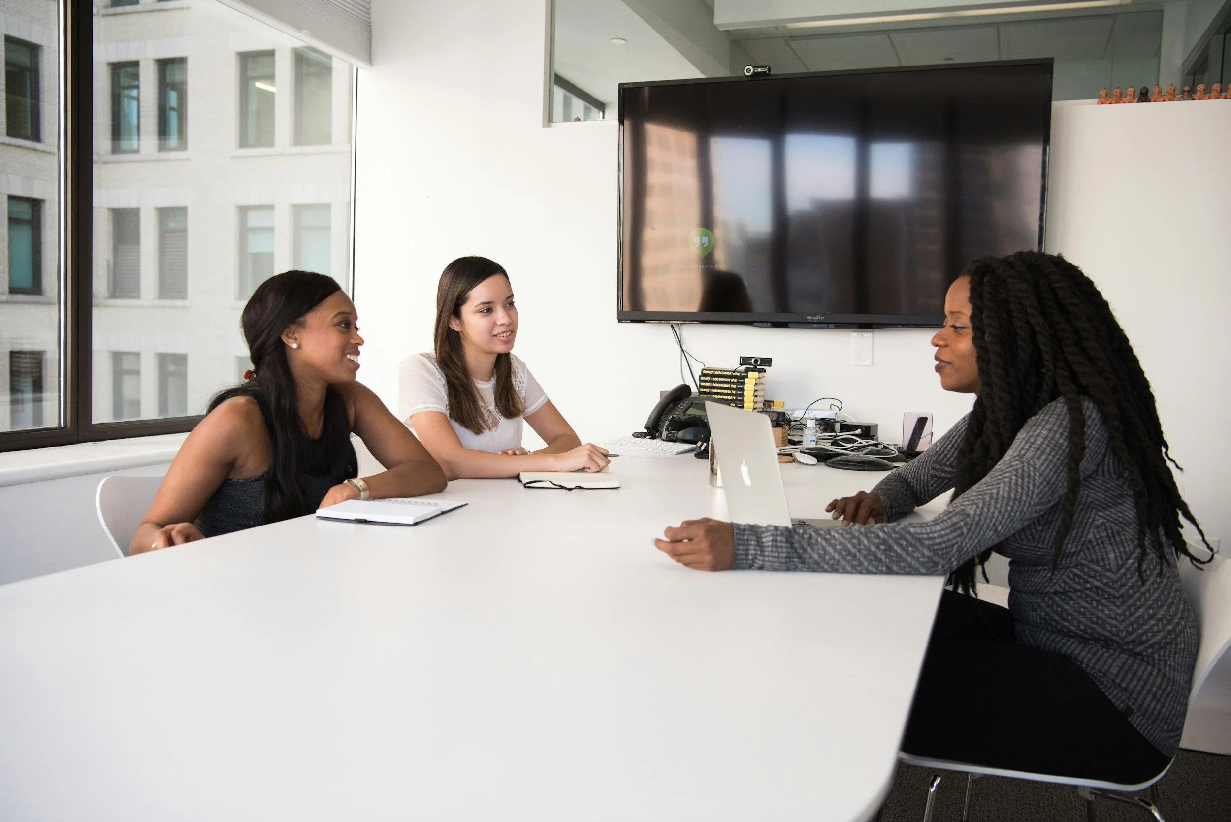 Two women sitting at a table talking. One Has a laptop and a grey sweater. The other has a notebook and a white tshirt.