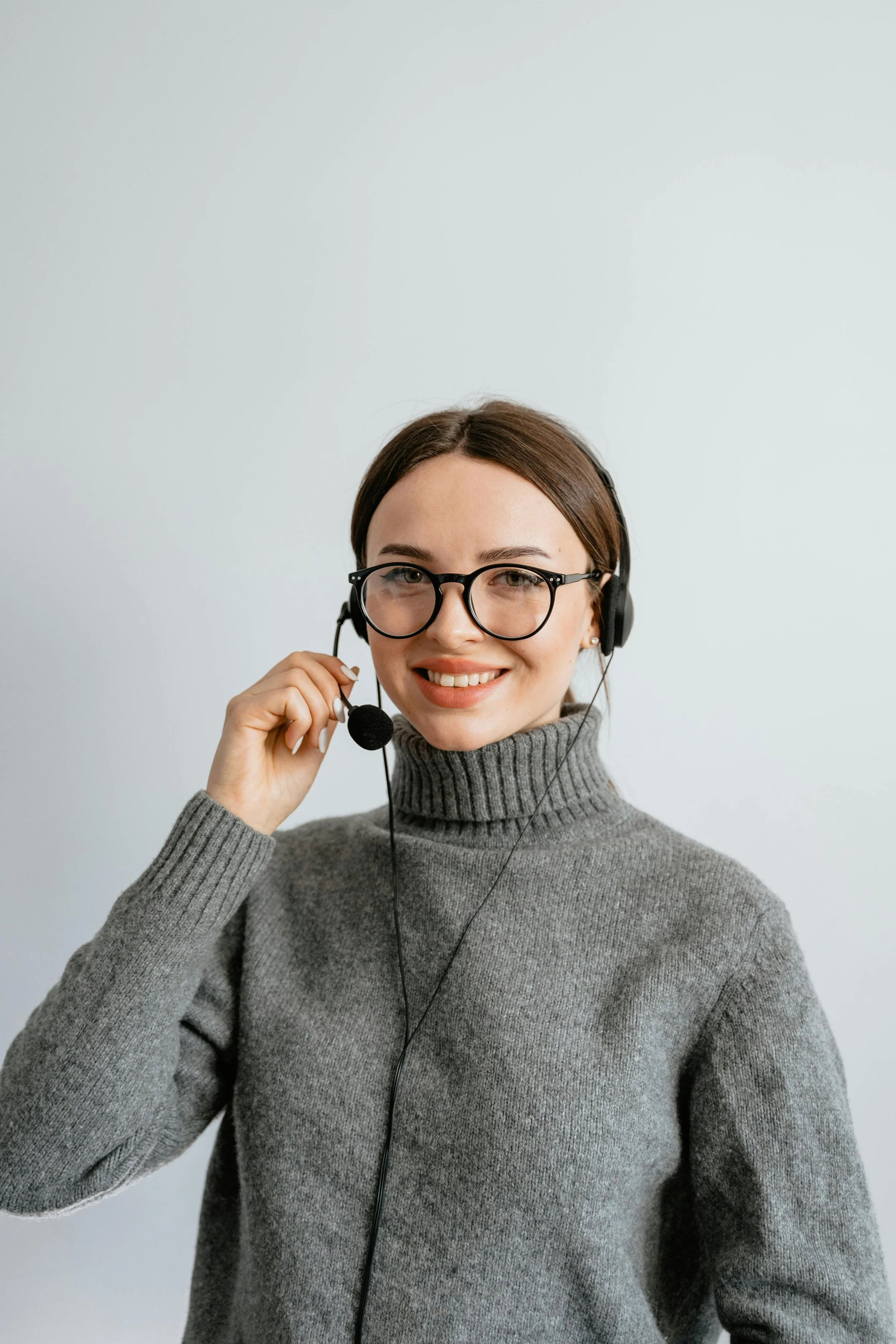 Woman wearing a headset and a grey turtleneck sweater and black rim glasses is smiling at the camera.