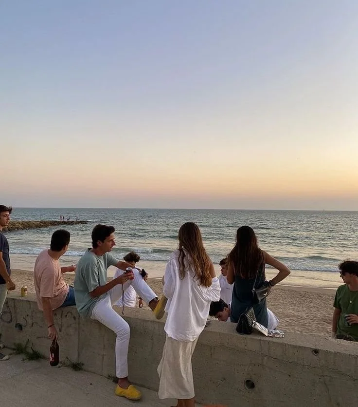 Group of young people at the beach during sunset, sitting and standing by a concrete ledge, with the ocean in the background.