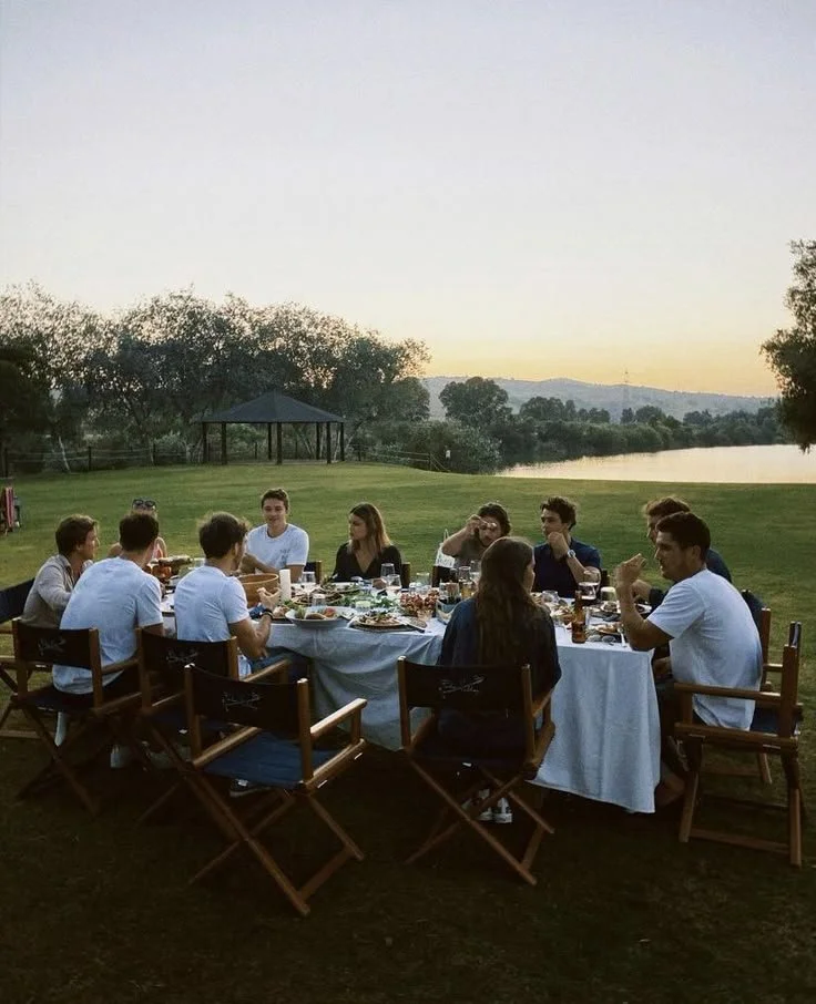 A group of people enjoying an outdoor dinner at a long table set with food and drinks, near a lake during sunset.