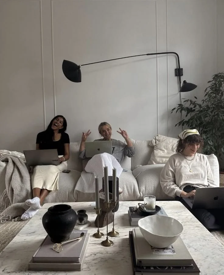 Three women sitting on a white sofa, using laptops, surrounded by a coffee table with decorative objects, candles, books, and bowls in a modern living room.