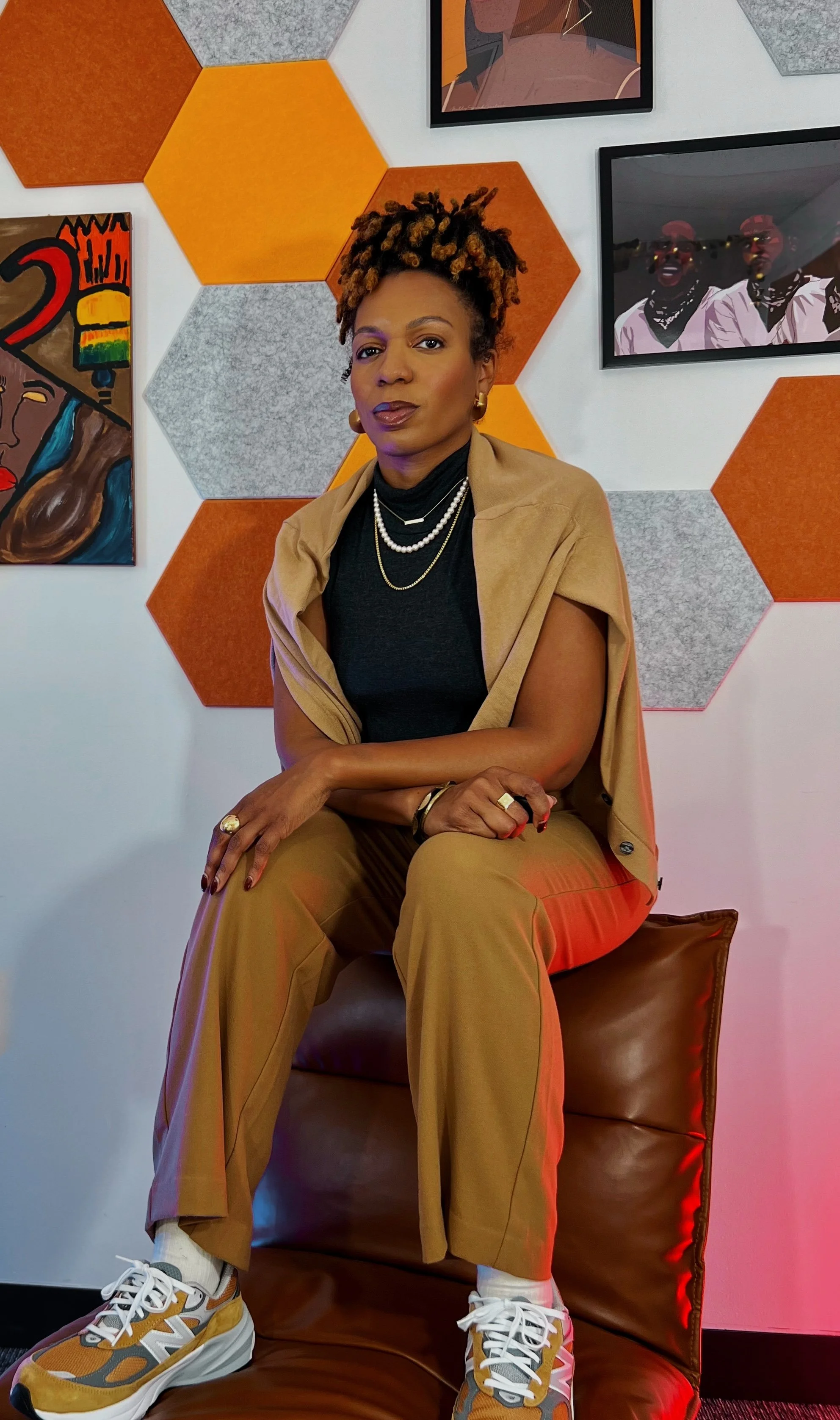 A woman with short dreadlocks sitting on a brown leather chair in front of a wall with orange and gray hexagonal panels and framed artwork, wearing a black top, beige pants, a beige jacket draped over her shoulders, and white sneakers with yellow accents, accessorized with layered necklaces, rings, earrings, and a bracelet.
