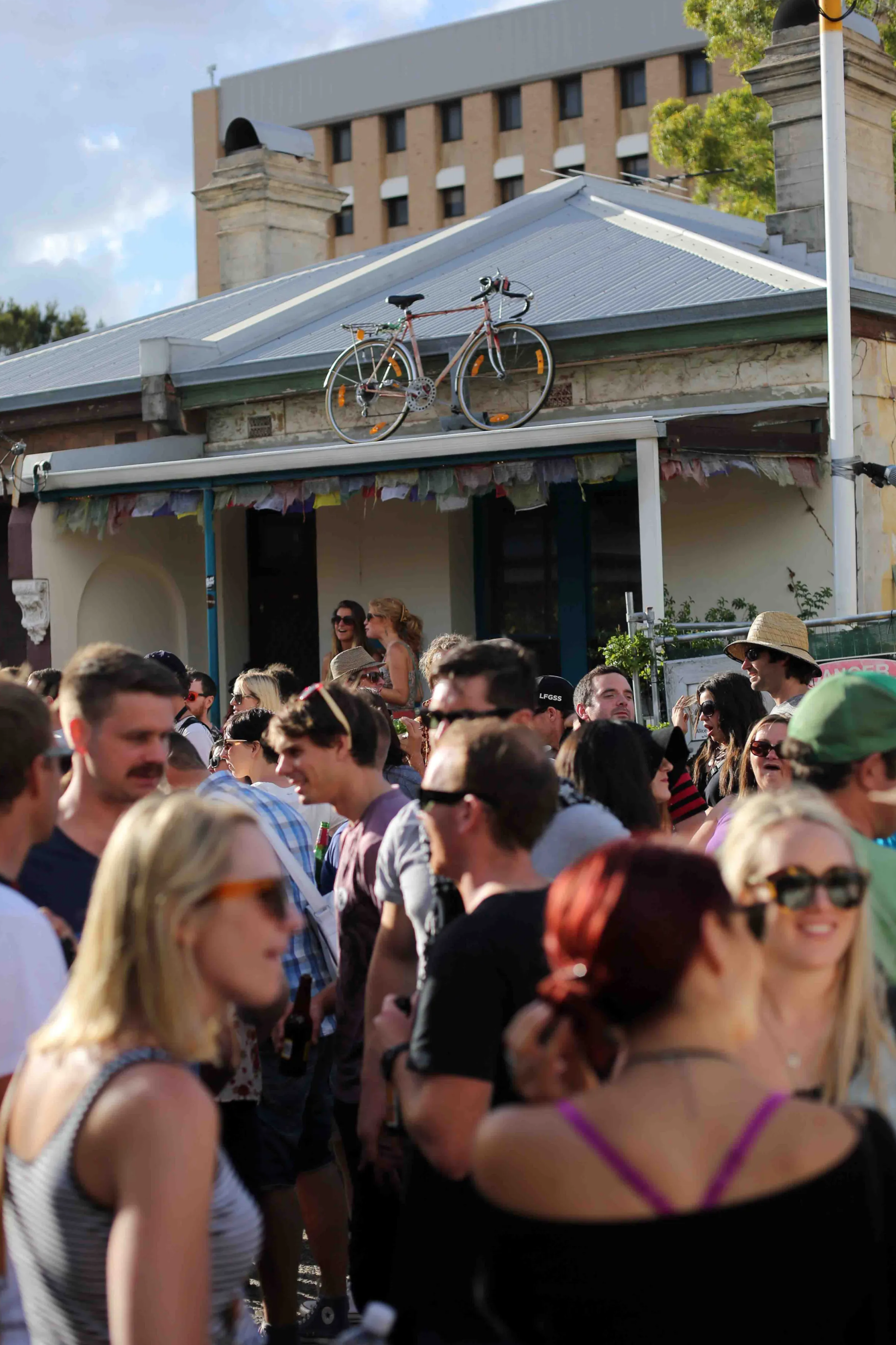 People gathered outdoors for a festival or event, with some wearing casual clothing and sunglasses, and a building with a bicycle on the roof in the background.