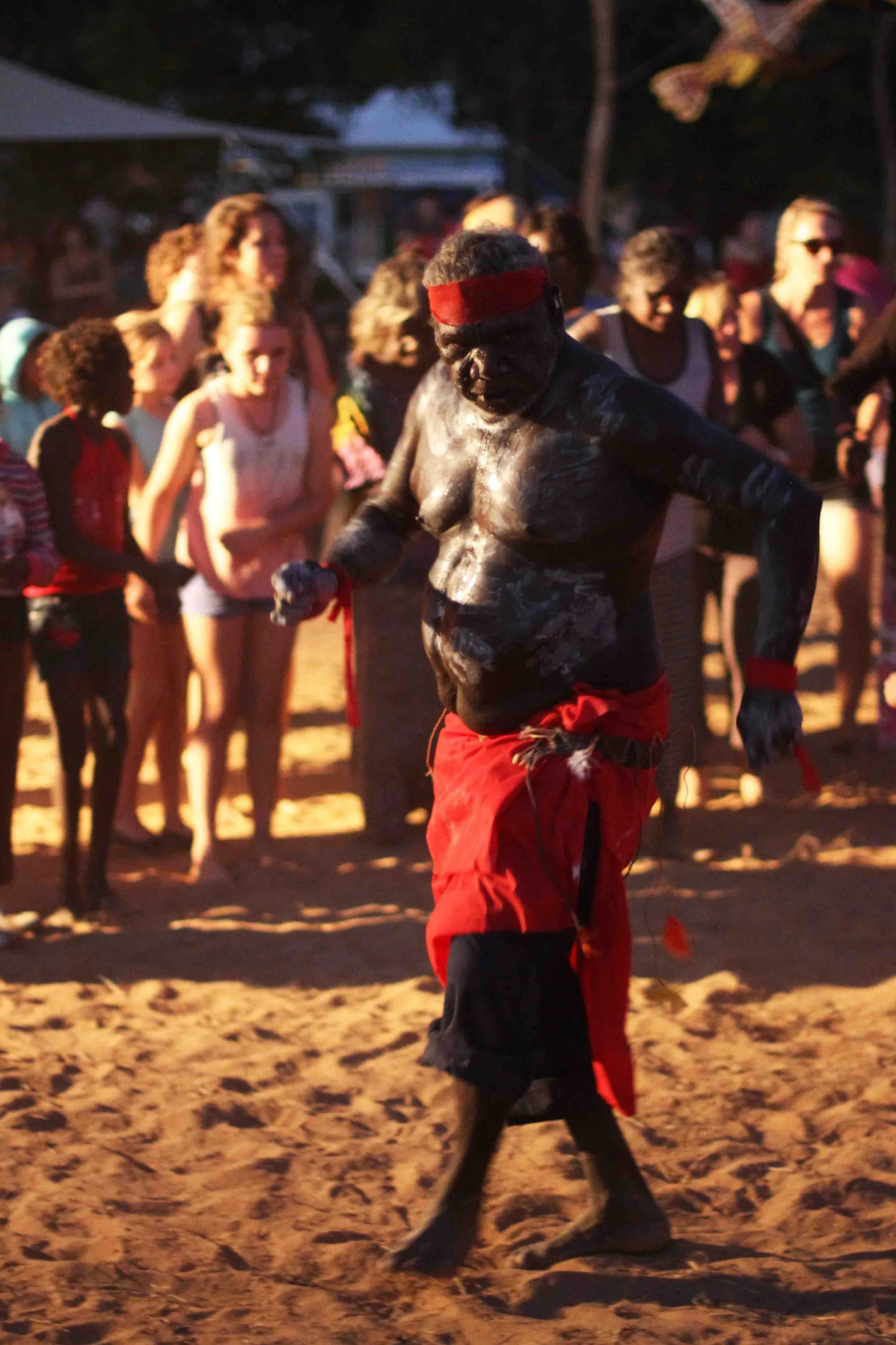 A traditional Baining fire dancer performing at night, covered in white paint, wearing a red loincloth, with an audience watching in the background.