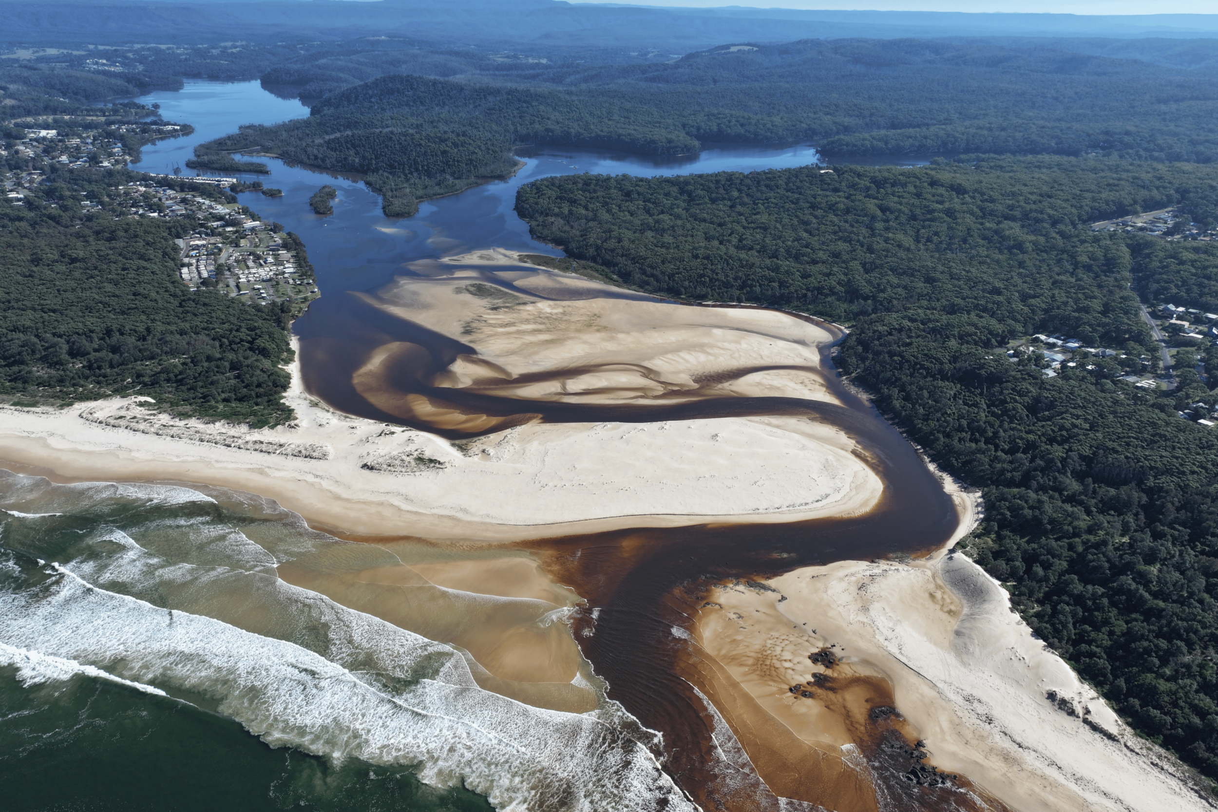 Aerial view of a beach with sand dunes and a river flowing into the ocean, surrounded by lush green forests and residential areas.