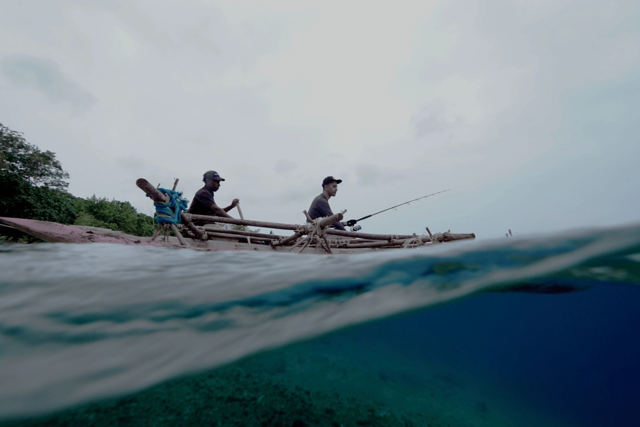 Two men fishing on a wooden boat near a forested shoreline under a cloudy sky, captured partly underwater.