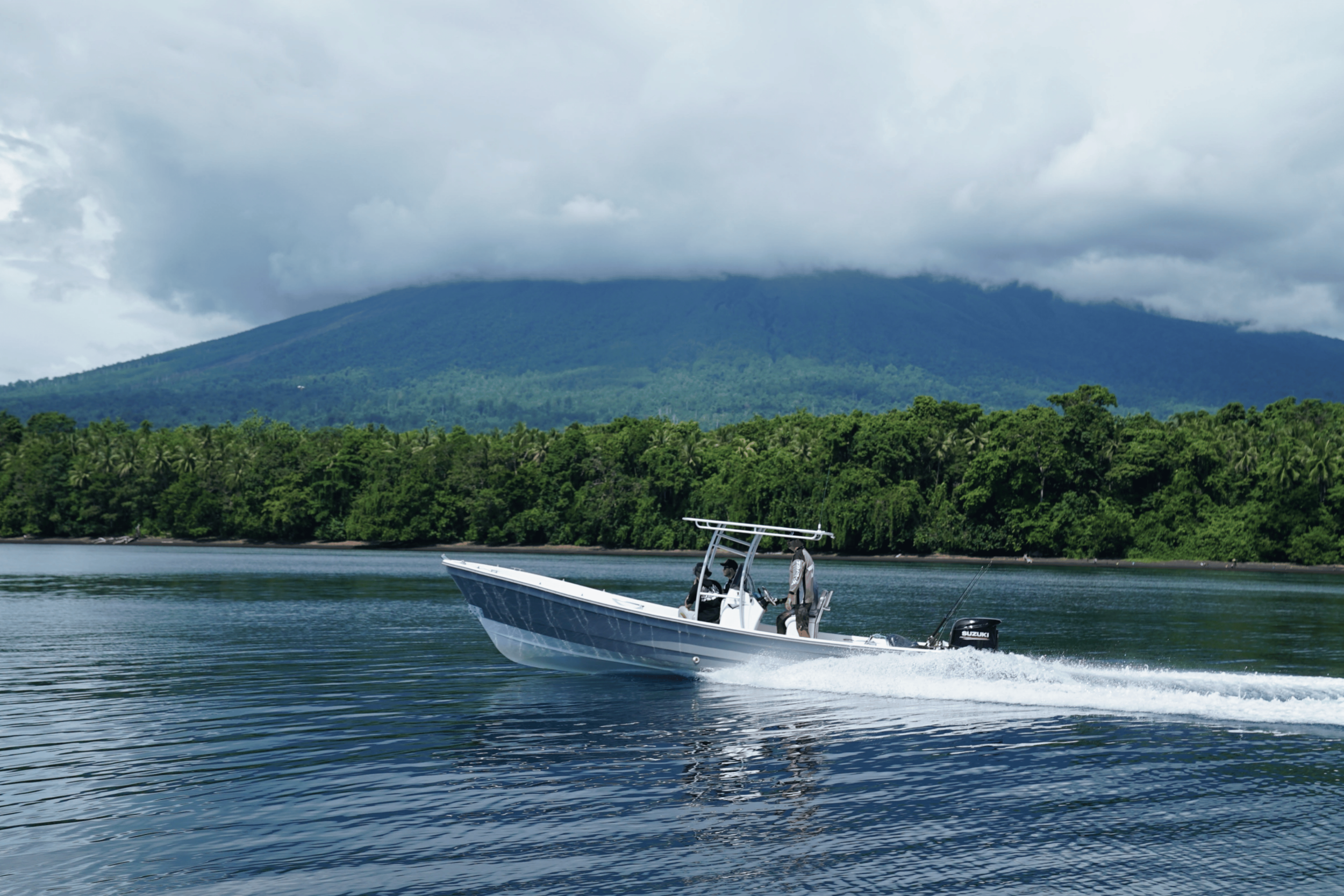 A motorboat with a person on board moving across a lake, with forested hills and a mountain covered in clouds in the background.