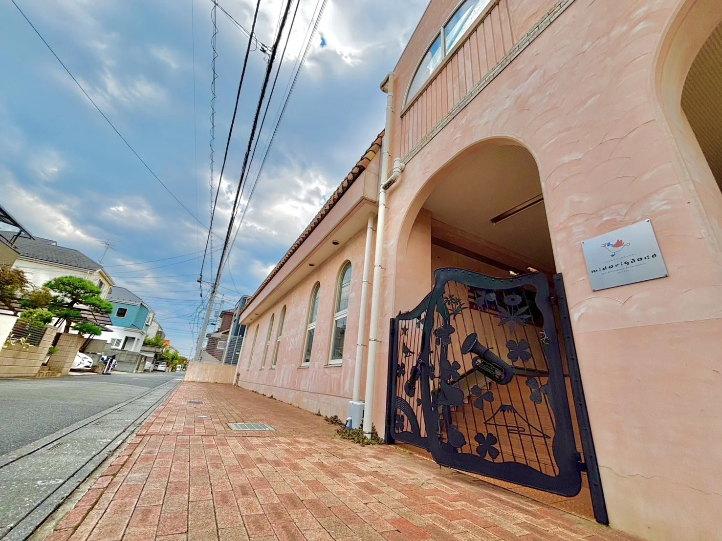 ピンク色の建物の入り口に装飾された黒いゲートがあり、サインには鳥のロゴと英語の文字が描かれている。通りにはレンガ舗装が続き、空は青空に少し雲が浮かんでいる。