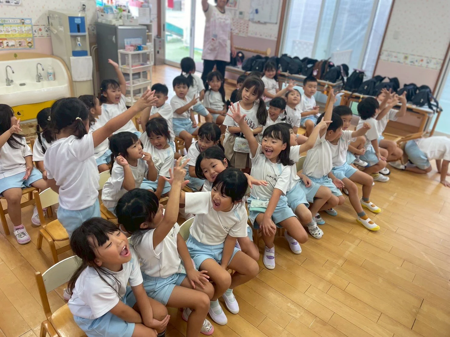 幼稚園の教室で子供たちが楽しそうに遊び、手を挙げたり笑ったりしています。