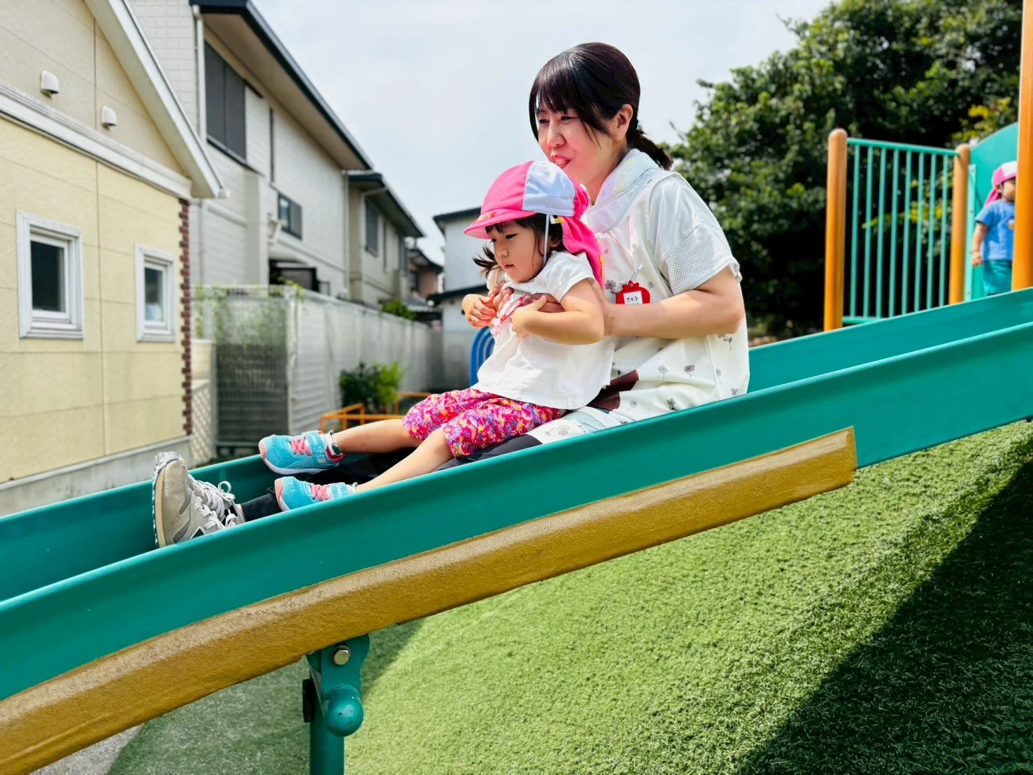 幼稚園児の女の子が滑り台を滑っている。女性の先生が背後から支えている。