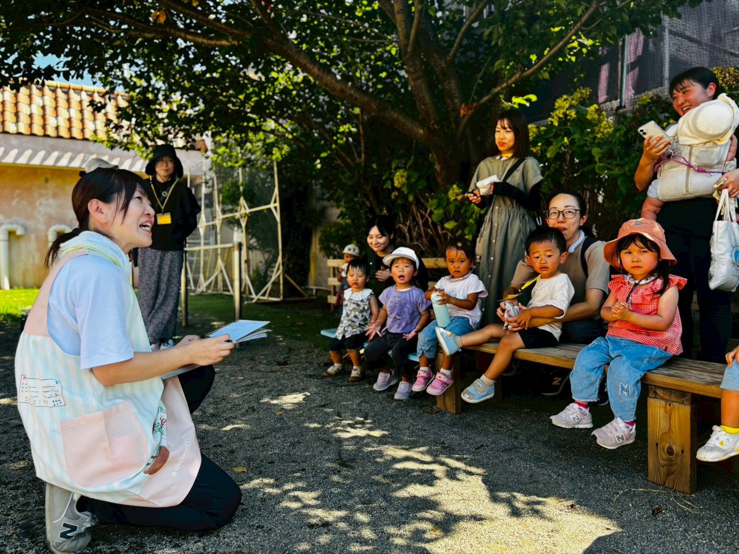 子供と大人たちが屋外の木やベンチの下で集まっている風景。子供たちが座り、大人たちが立っている。誰かが話していて、子供たちが興味を持って見ている。