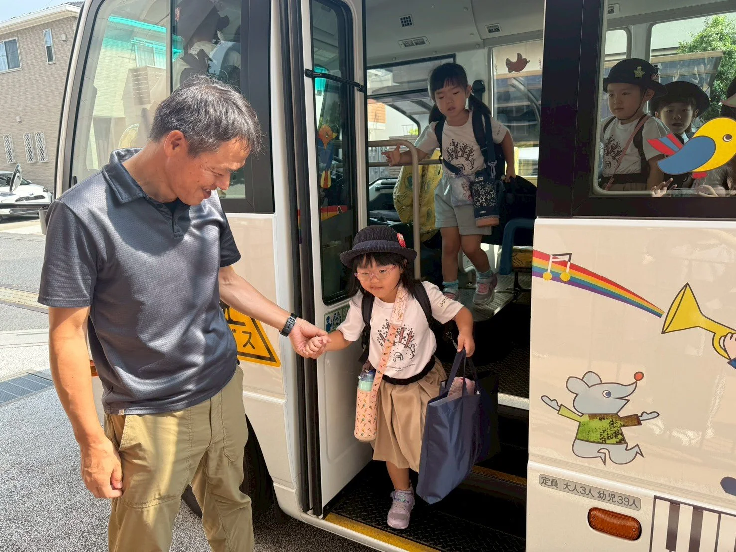男性がバスから降りる子供を手で支えながら見守る場面。子供たちはバスから降りている途中で、学校の制服を着ている。