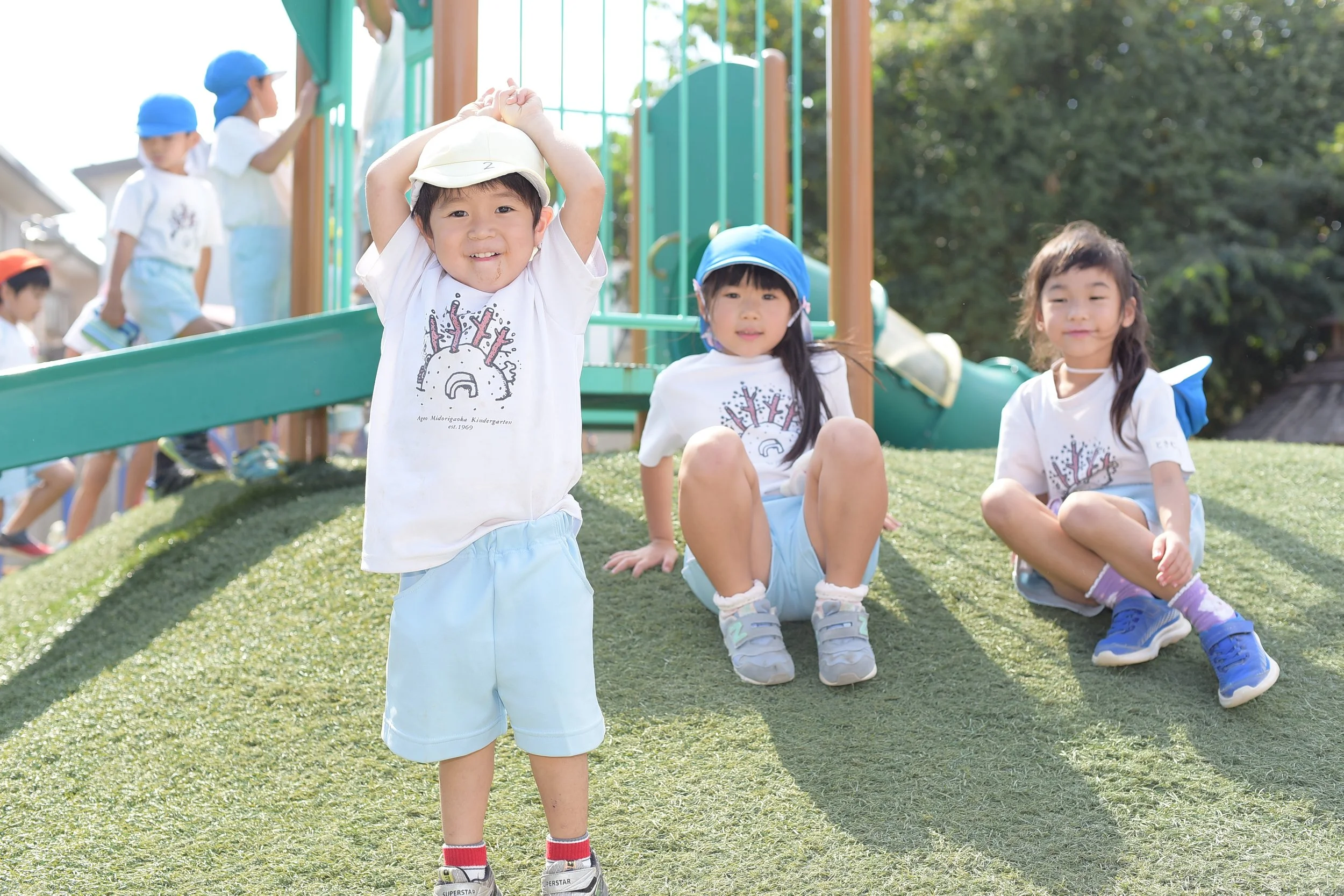 公園で遊ぶ幼稚園児たち。1人の男の子が帽子をかぶりながら笑顔で両手を頭の上に置いています。2人の女の子は座っており、どちらも青の帽子と白いTシャツを着ています。背景には滑り台や木が見えます。