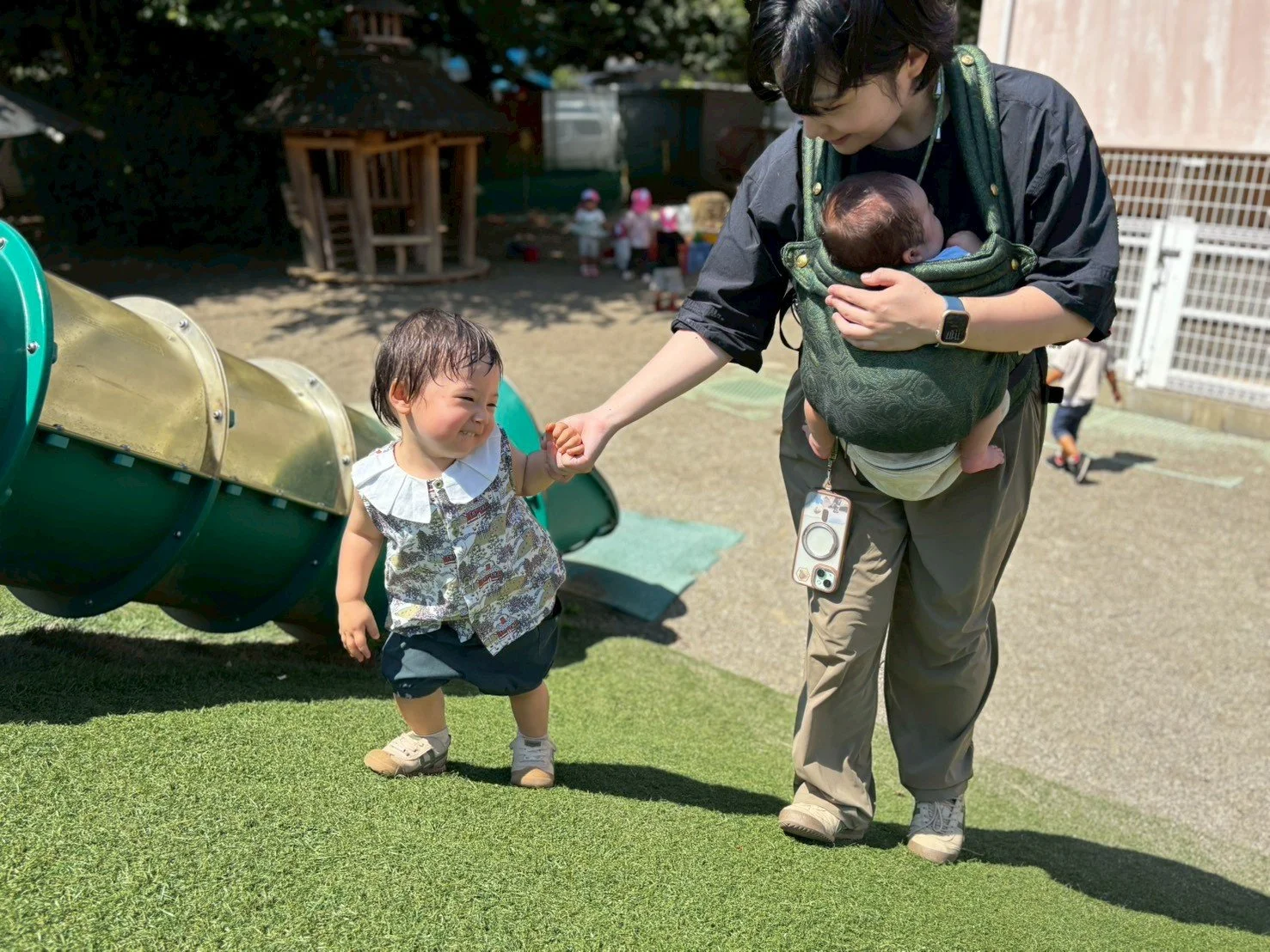子供と母親が遊び場で遊んでいる。母親は赤ちゃんを抱きながら子供の手をつなぎ、子供は笑顔で母親に手を引かれている。静かな公園の遊具と背景の子供たちが見える。