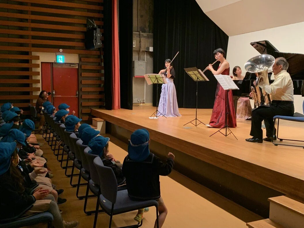 コンサートホールで小学生の子供たちが椅子に座って音楽会を見ている。舞台には5人の弦楽器と管楽器の演奏者が演奏している様子が見える。