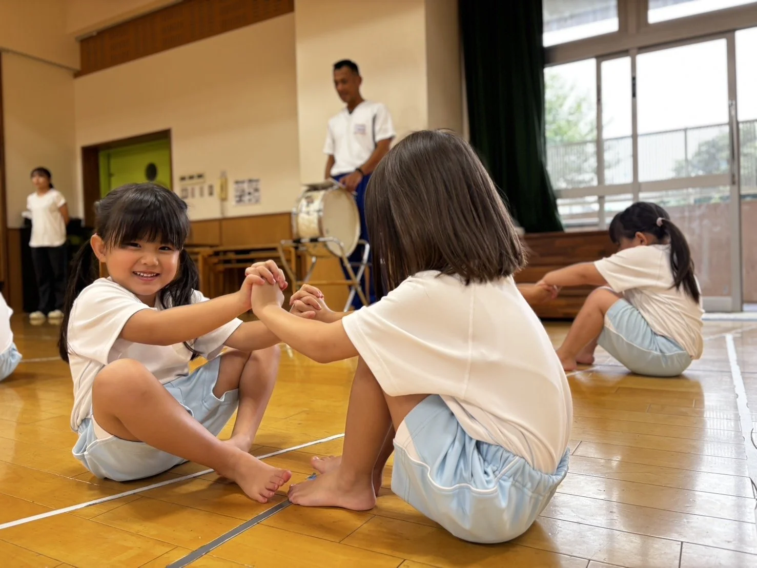 木曜日の幼稚園の室内で、2人の女の子が手をつないで遊んでいる。背景には別の子供や音楽を演奏する男性がいる。