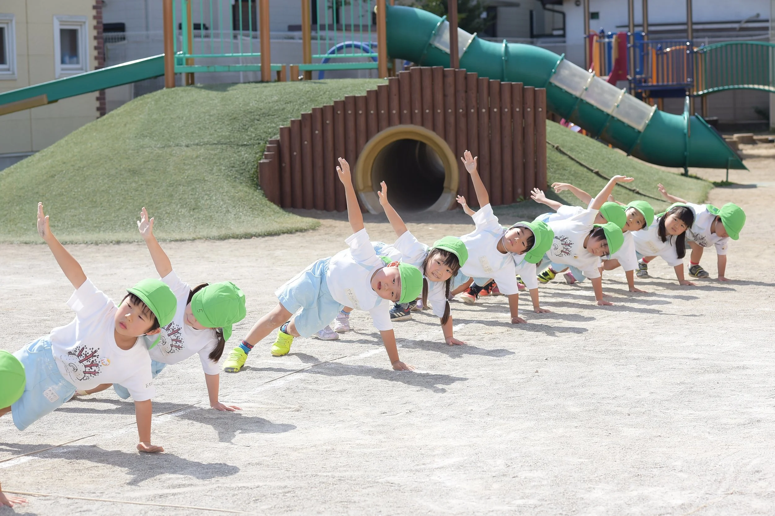 幼稚園児が外の遊び場で片手と片足をついてバランスを取りながら横になっている順番に並んでいる。