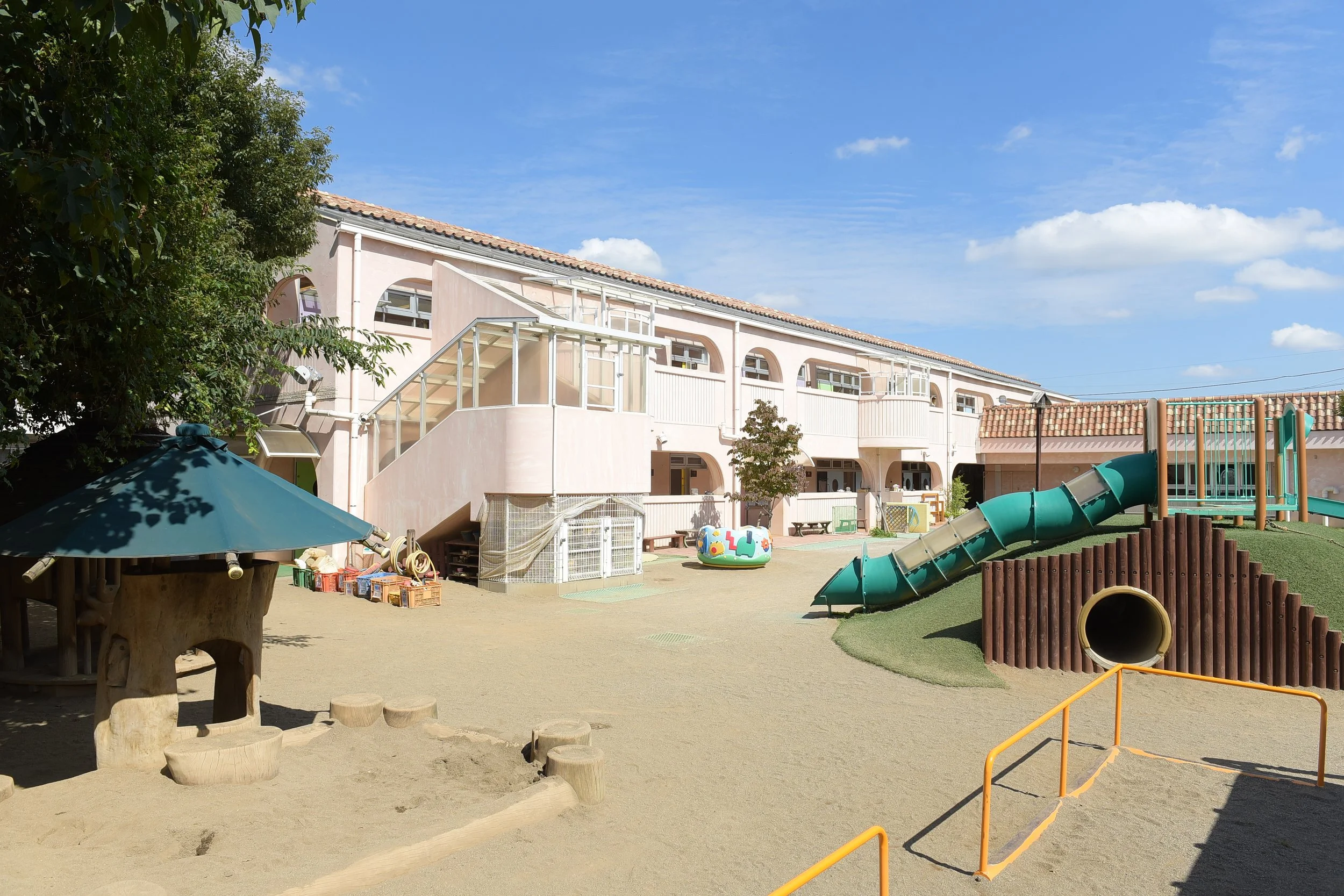 幼稚園の遊び場で、滑り台や砂場といった遊具がある屋外のプレイエリア。背景にはピンク色の建物と青空が見える。