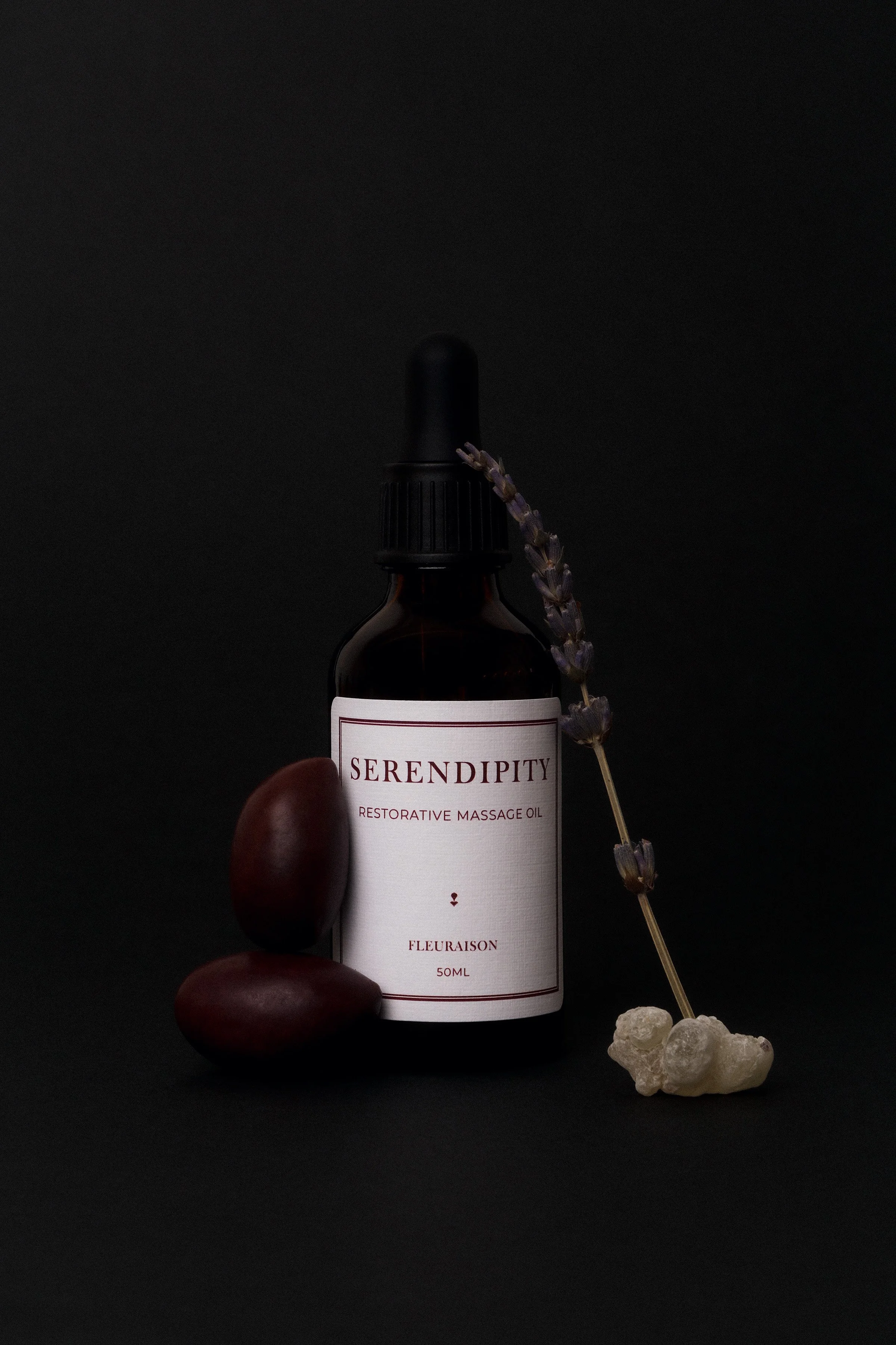 Massage oil bottle styled with olives, lavender and frankincense resin against a black backdrop