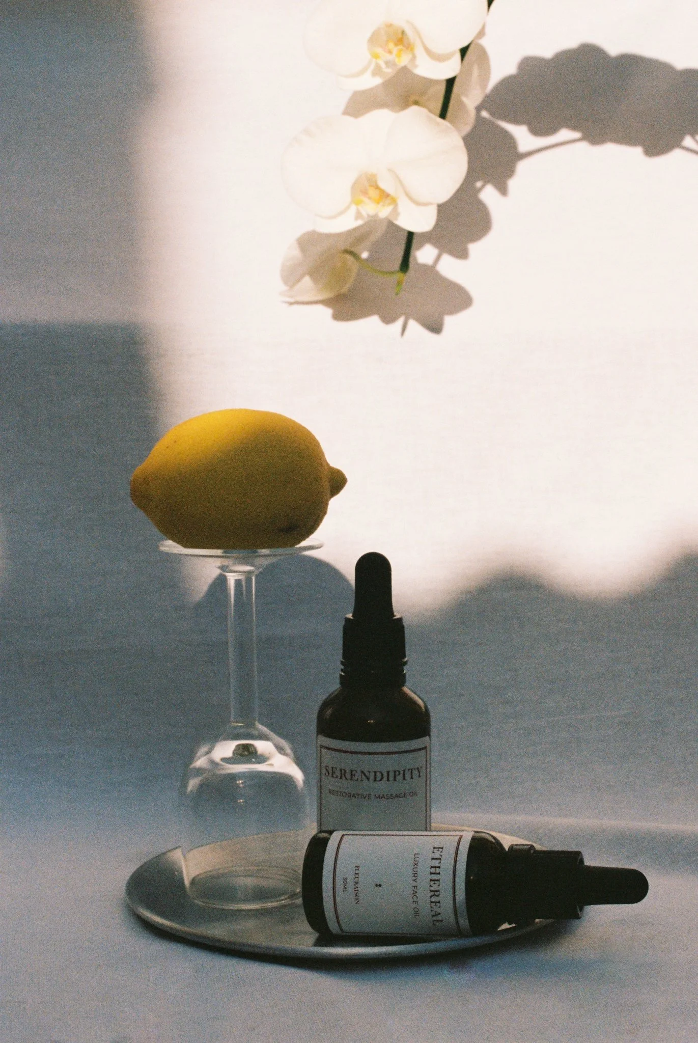 Face oils labelled Serendipity and Ethereal on a silver tray, on a white surface with soft lighting, styled with white orchids and a lemon on a wine glass.