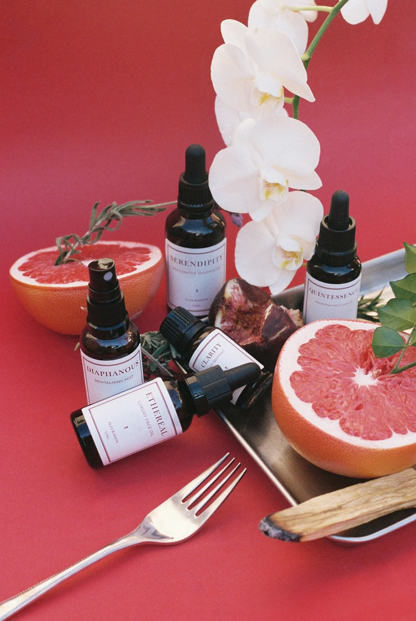 A flat lay of skincare bottles labelled Serendipity, Quintessence, Diaphanous, Clarity and Ethereal styled with sliced grapefruit, white orchids, rosemary, palo santo, a fig and a silver fork against a bright red background.