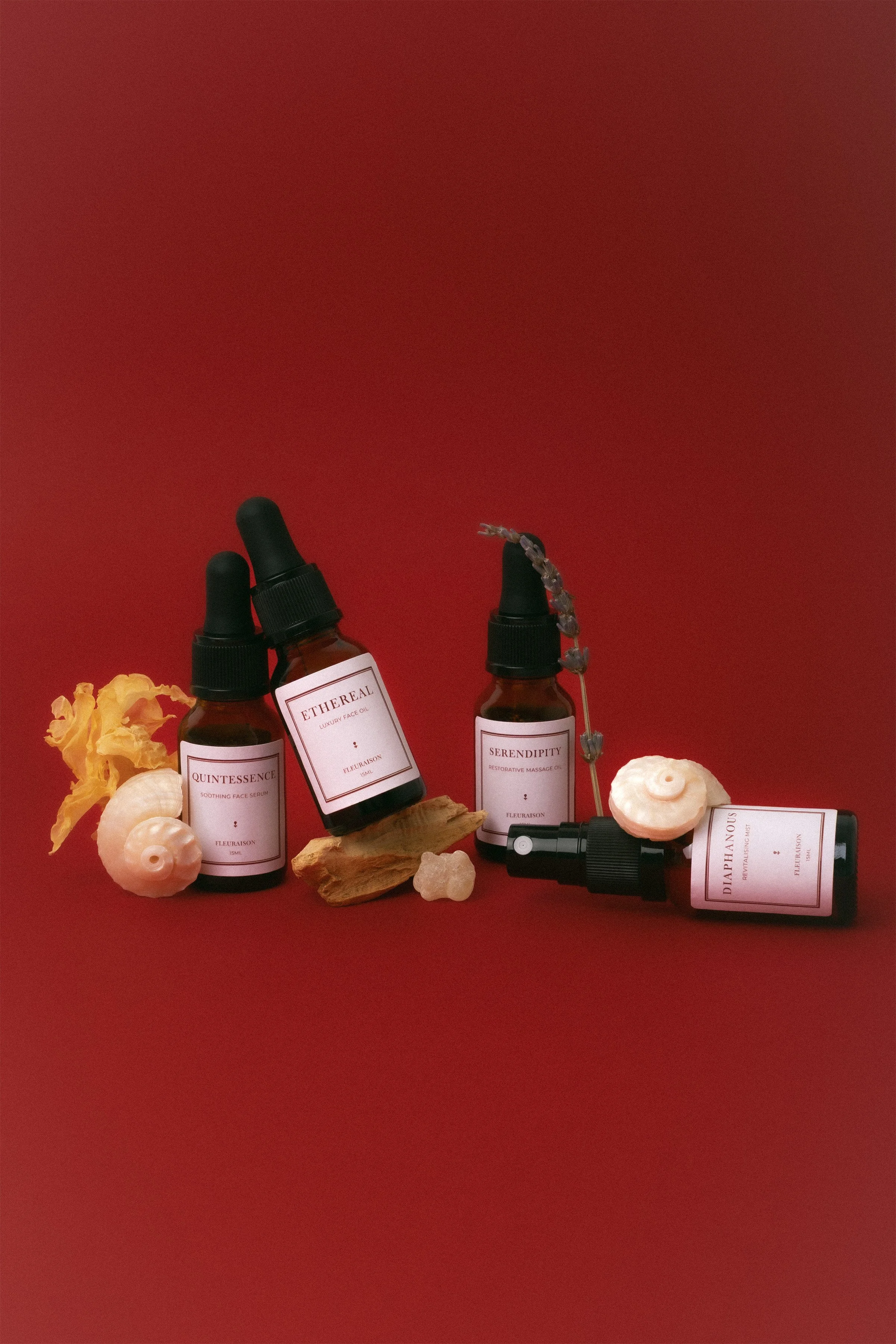 Fleuraison mini travel bottles of Quintessence Face Serum, Ethereal Face Oil, Serendipity Massage Oil and Diaphanous Mist, styled with shells, sandalwood, snow mushroom against a dark red background.