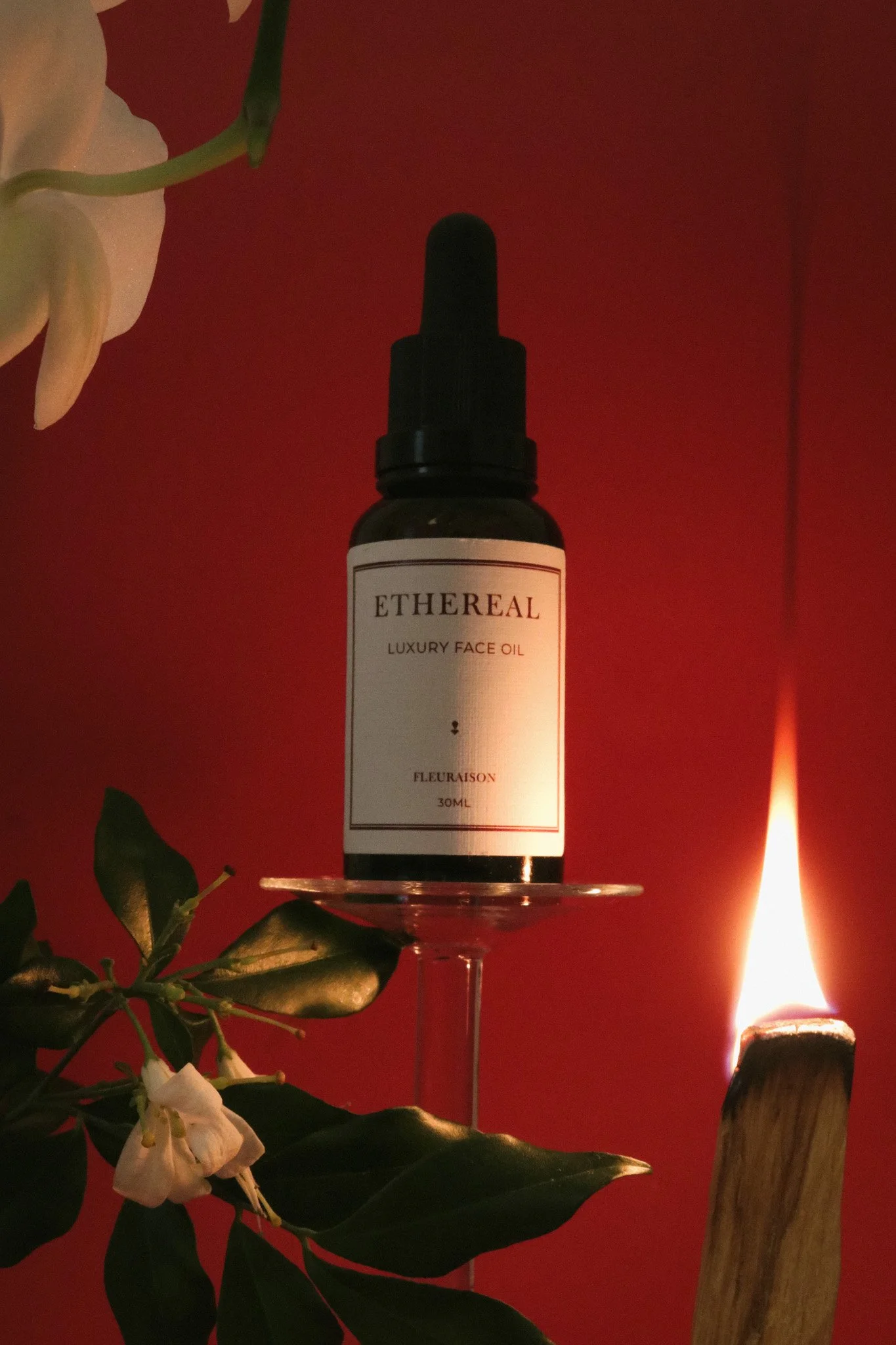 Fleuraison Ethereal Luxury Face Oil displayed on an upside down wineglass, styled with a lit palo santo stick and green and white botanicals against a red background.