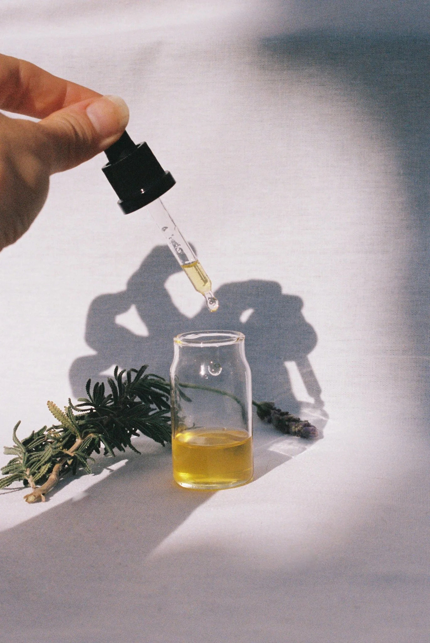 Yellow face oil in a glass container with a hand dispensing the oil from a dropper, styled with rosemary and lavender.