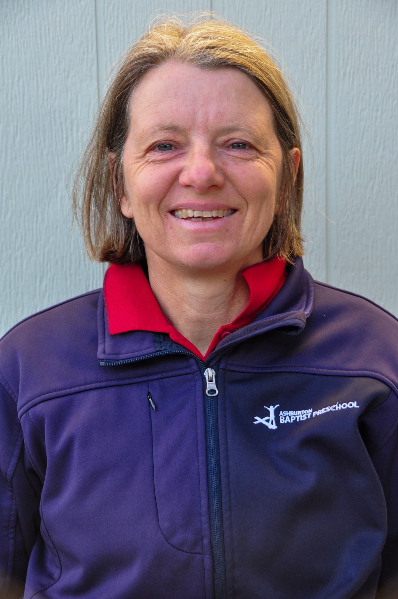 Portrait of a woman with shoulder-length brown hair, wearing a red polo shirt with a logo that reads 'Ashland Baptist School,' sitting on a wooden surface against a light-colored wall.