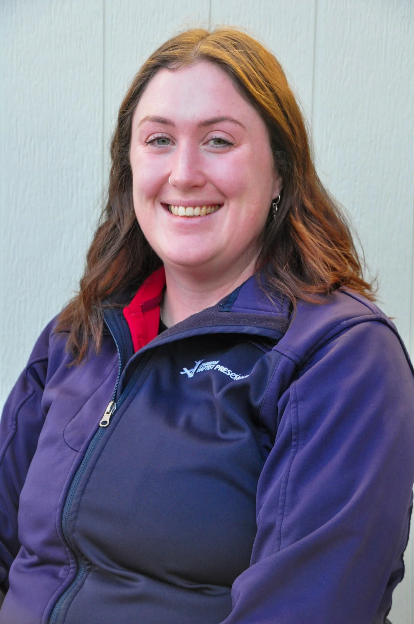 A woman with tattoos on her arm, wearing a red polo shirt with 'Ashburton Baptist Preschool' logo, leaning on a black railing outdoors on a sunny day.