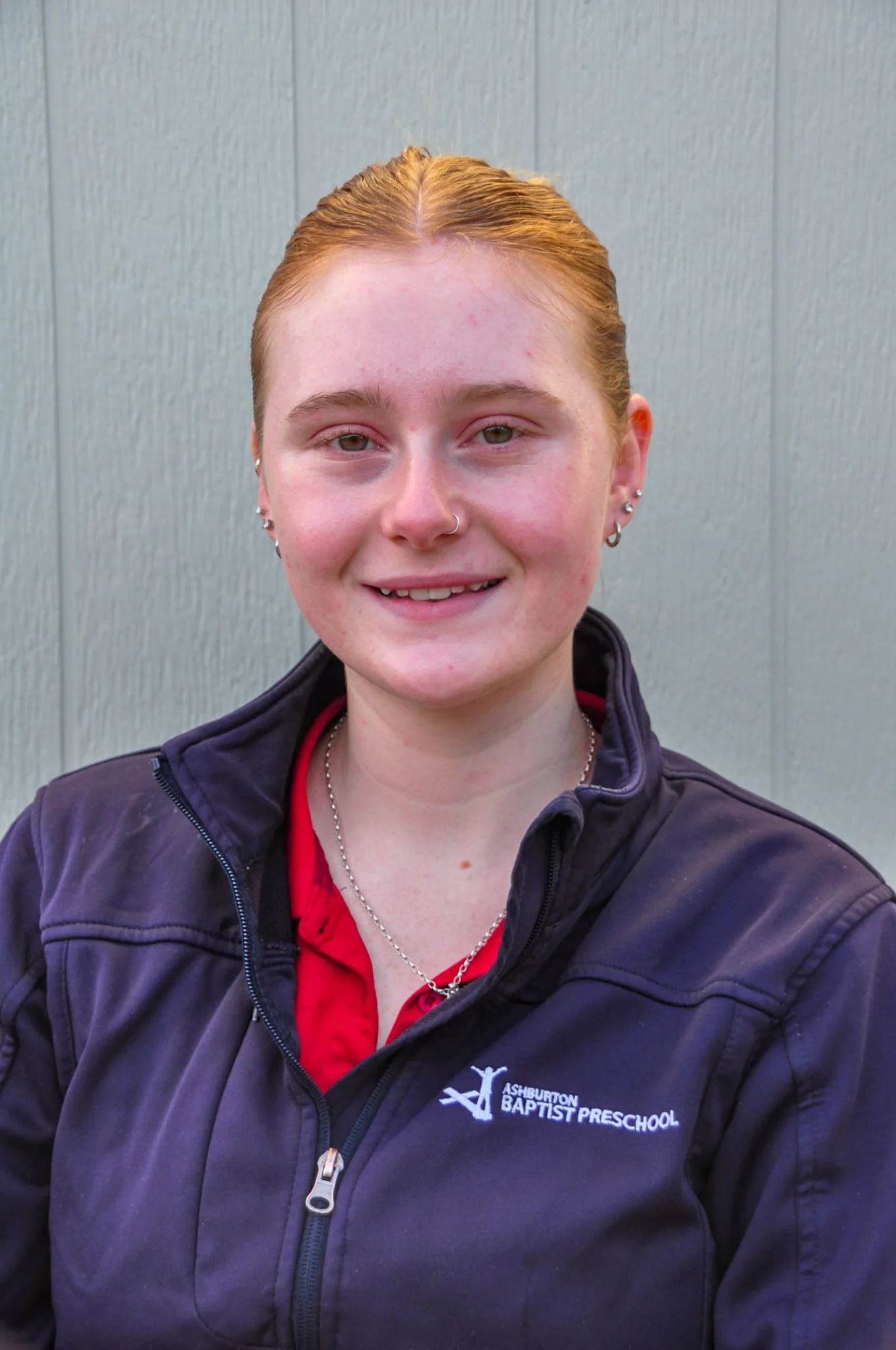 Young woman with brown hair in a ponytail, wearing a navy sweatshirt with 'Ashburton Baptist Preschool' logo, smiling with arms crossed outdoors near a black railing.