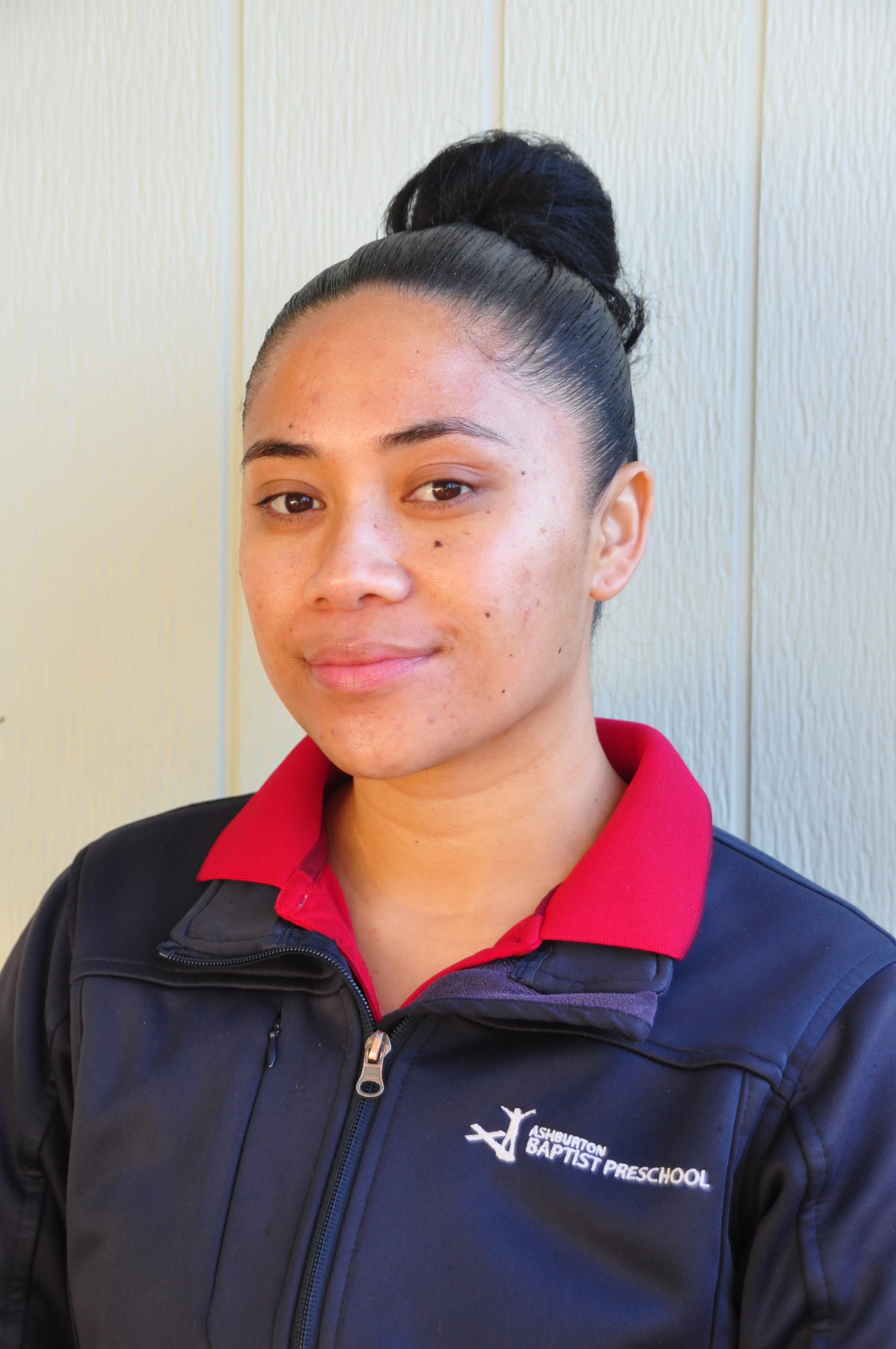 A woman with glasses and earrings, wearing a red polo shirt with a logo that reads 'Bentonville Baptist Preschool,' stands outdoors on a sunny day, leaning on a black railing with her hands clasped.