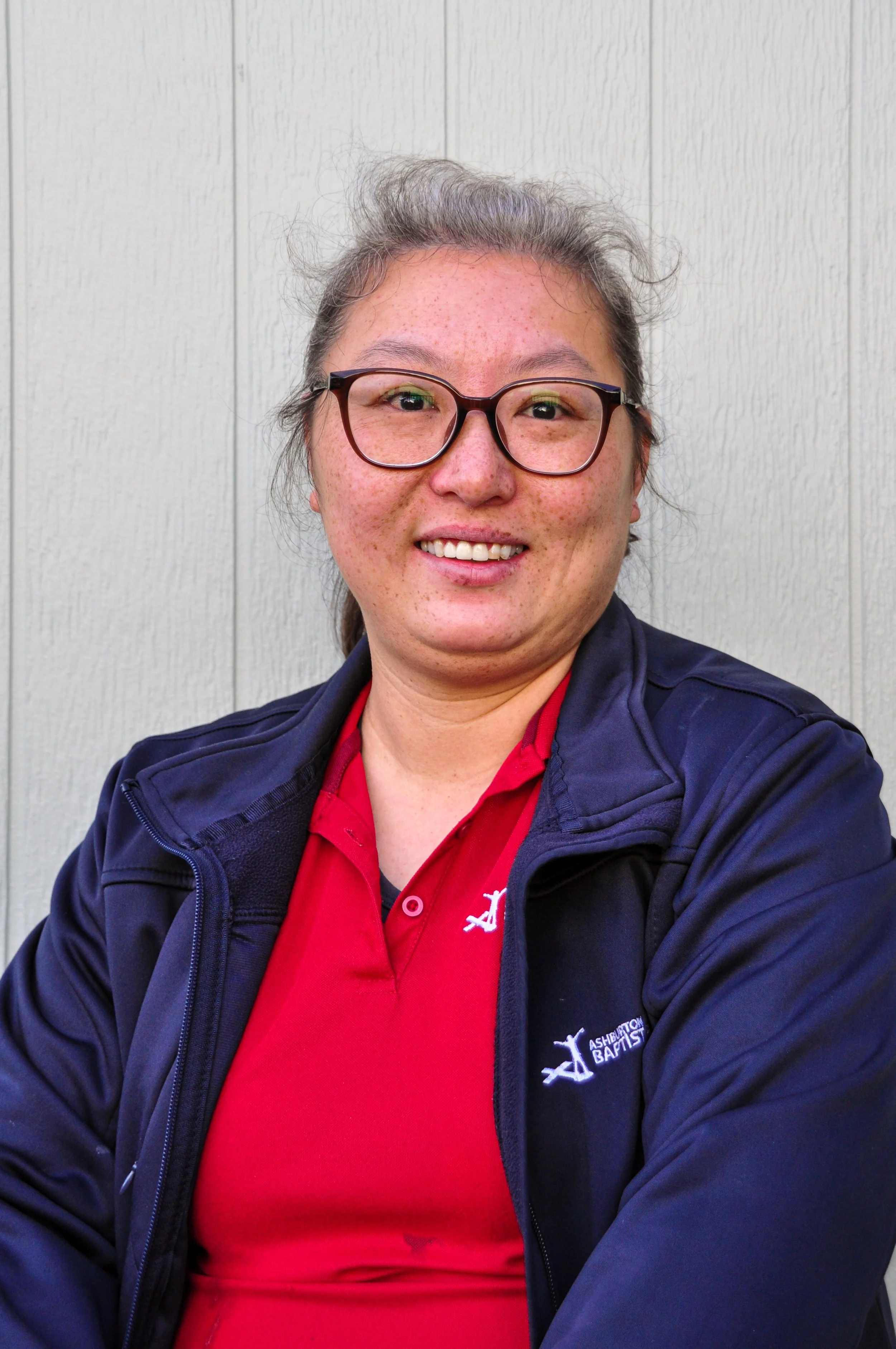 A woman with glasses and long hair leaning on a black railing outdoors, wearing a red shirt with 'Ashburton Baptist Preschool' logo, and a green long sleeve shirt underneath.