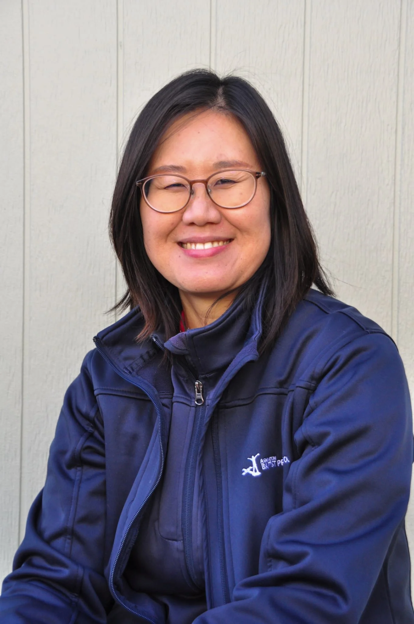 A woman with glasses and shoulder-length hair, wearing a sporty vest, leaning on a railing outdoors with a slight smile.