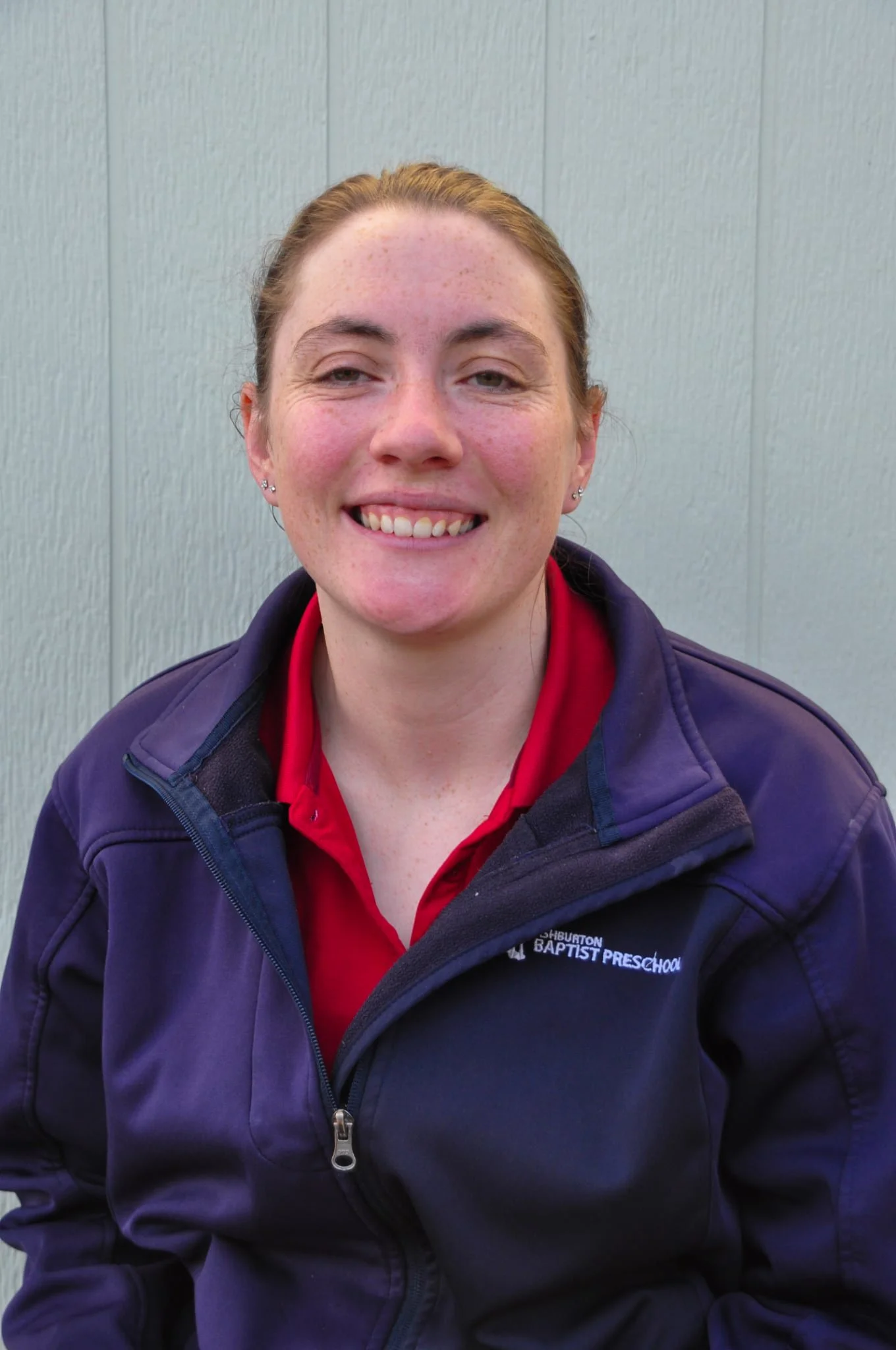 A woman with blonde hair smiling outdoors, wearing a red polo shirt with 'Ashburton Baptist Preschool' logo, leaning on a black and white fence, with a sunny sky and a covered area in the background.