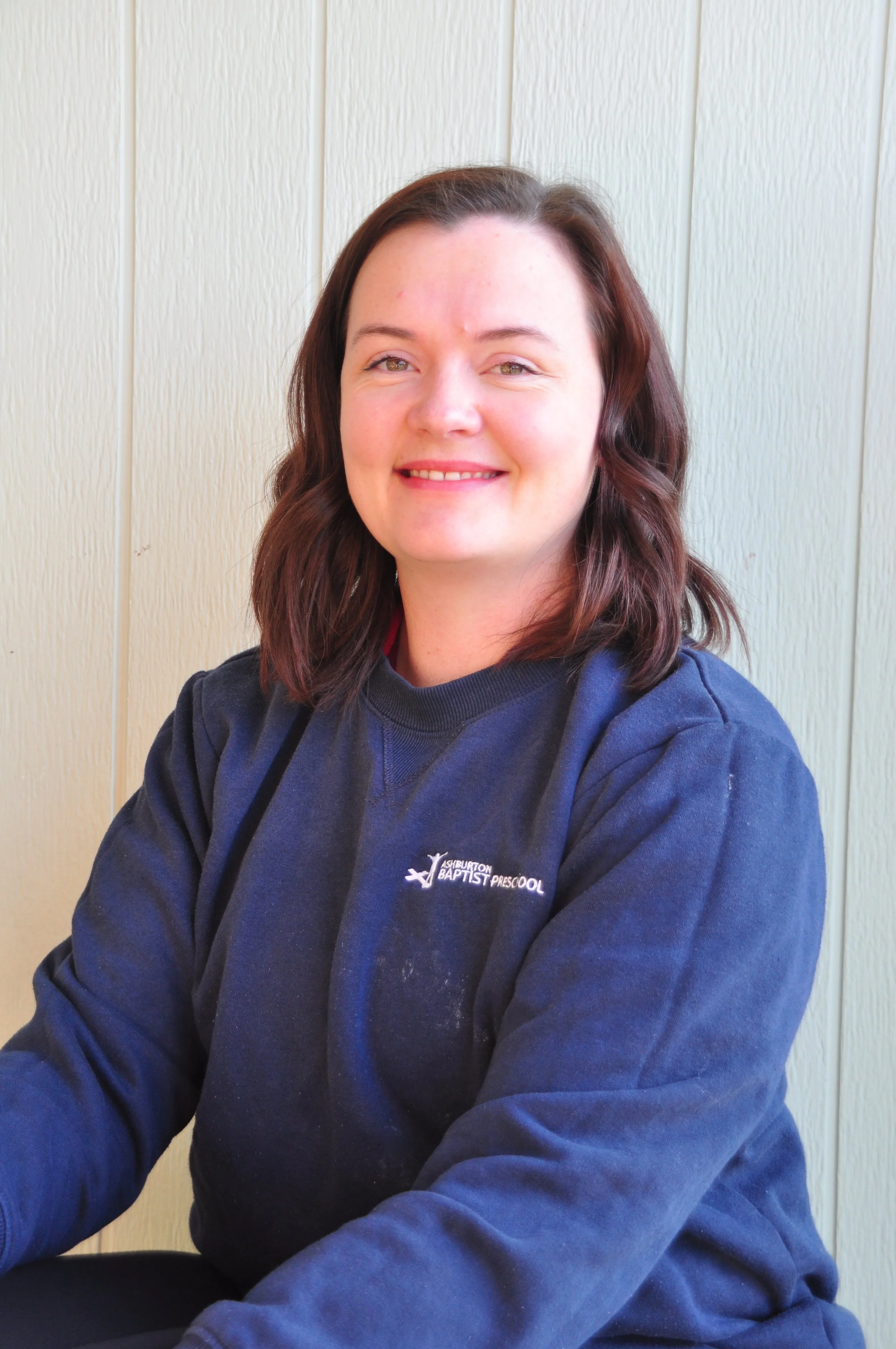 A woman with red hair wearing a red sports shirt, leaning on a black metal fence outdoors, smiling, with a pale sky and a blurred house in the background.