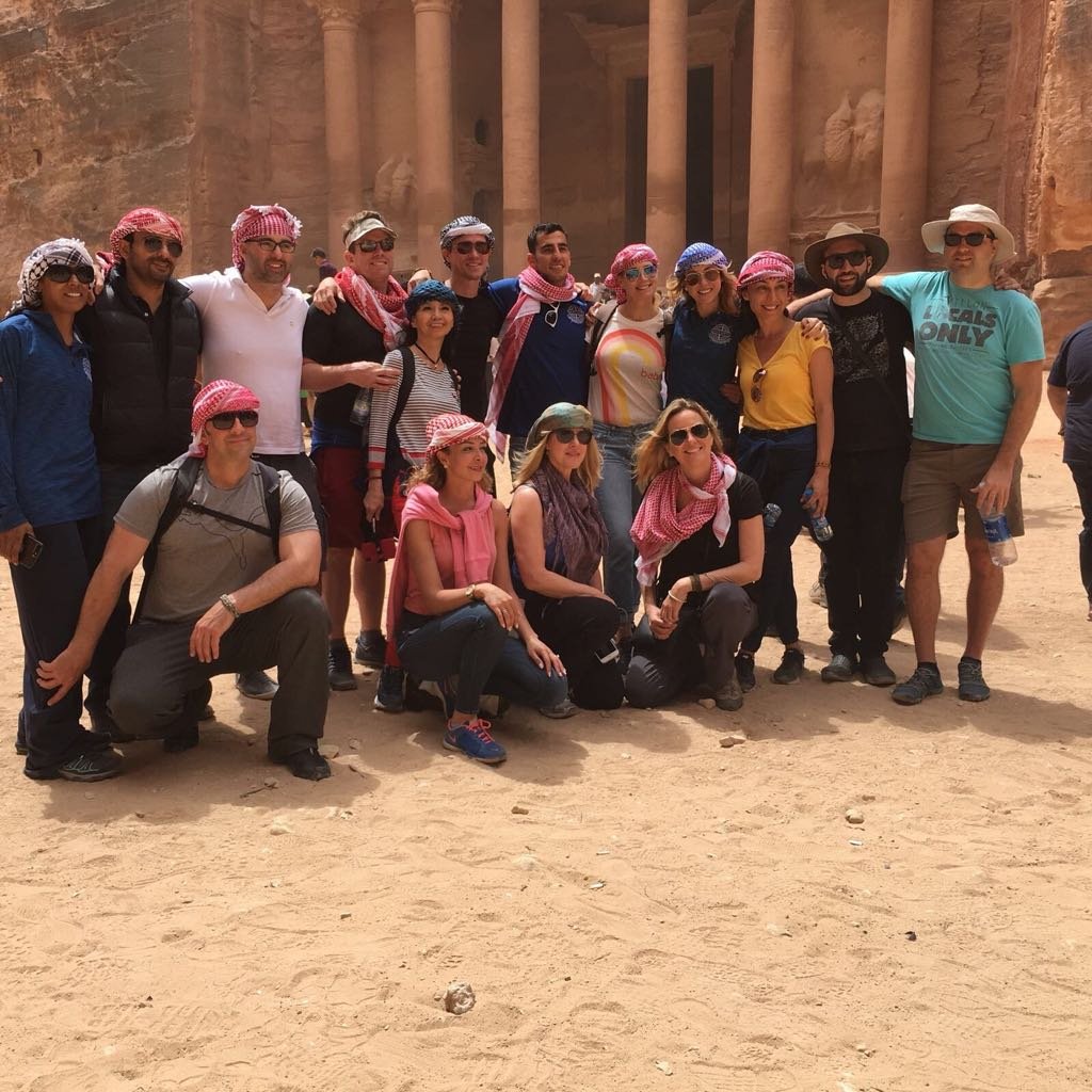 Group of tourists posing in front of ancient ruins, likely Petra, with sandy ground and carved stone walls in the background.