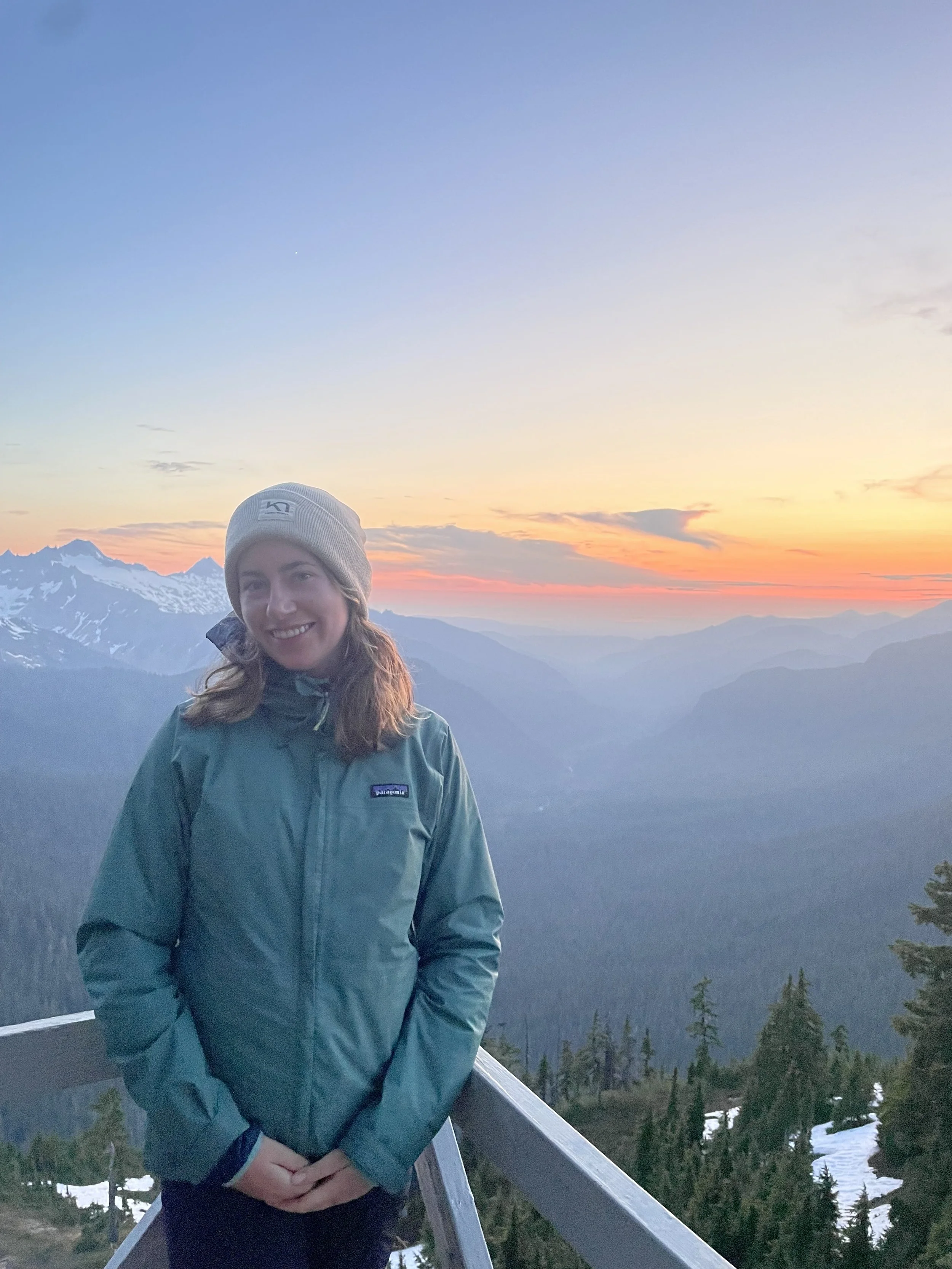 A woman in a teal Patagonia jacket and gray knit hat standing on a mountain overlook at sunset, with snow-capped peaks and a forested valley in the background.