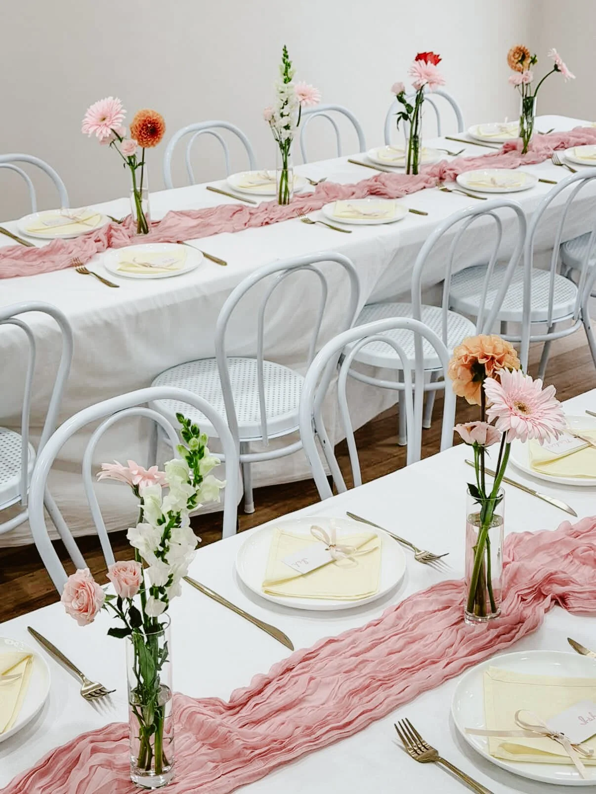 Long dining table decorated with pink fabric runner, tall vases of pink and white flowers, place settings with plates, napkins, and cutlery, white chairs.