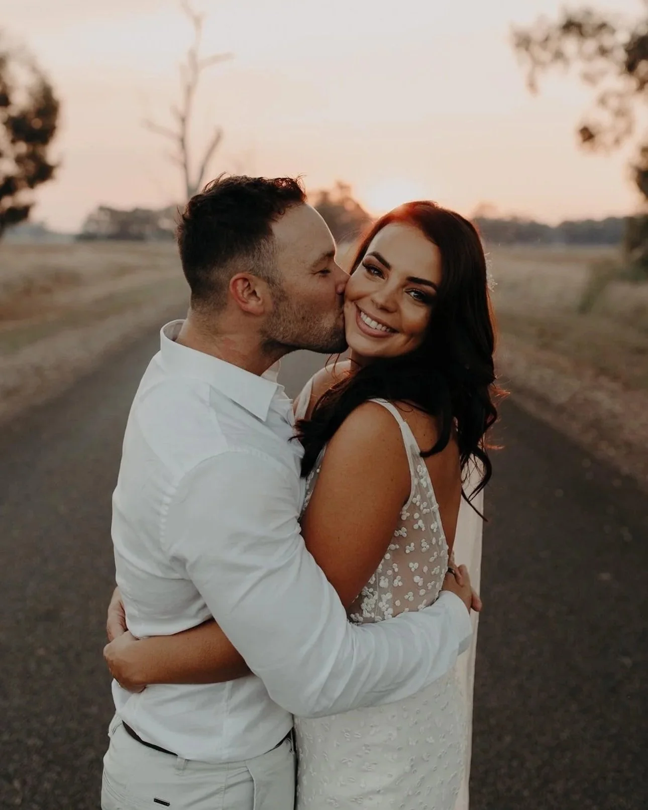 A man and woman embracing outdoors during sunset, with the man giving the woman a kiss on the cheek while she smiles at the camera.