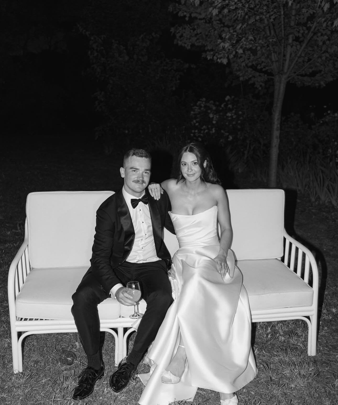 A black and white photo of a man and woman sitting on a bench outdoors at night, dressed in formal attire. The man is wearing a tuxedo and holding a glass of wine, and the woman is in a strapless gown. They are smiling, with the woman resting her arm on the man's shoulder.