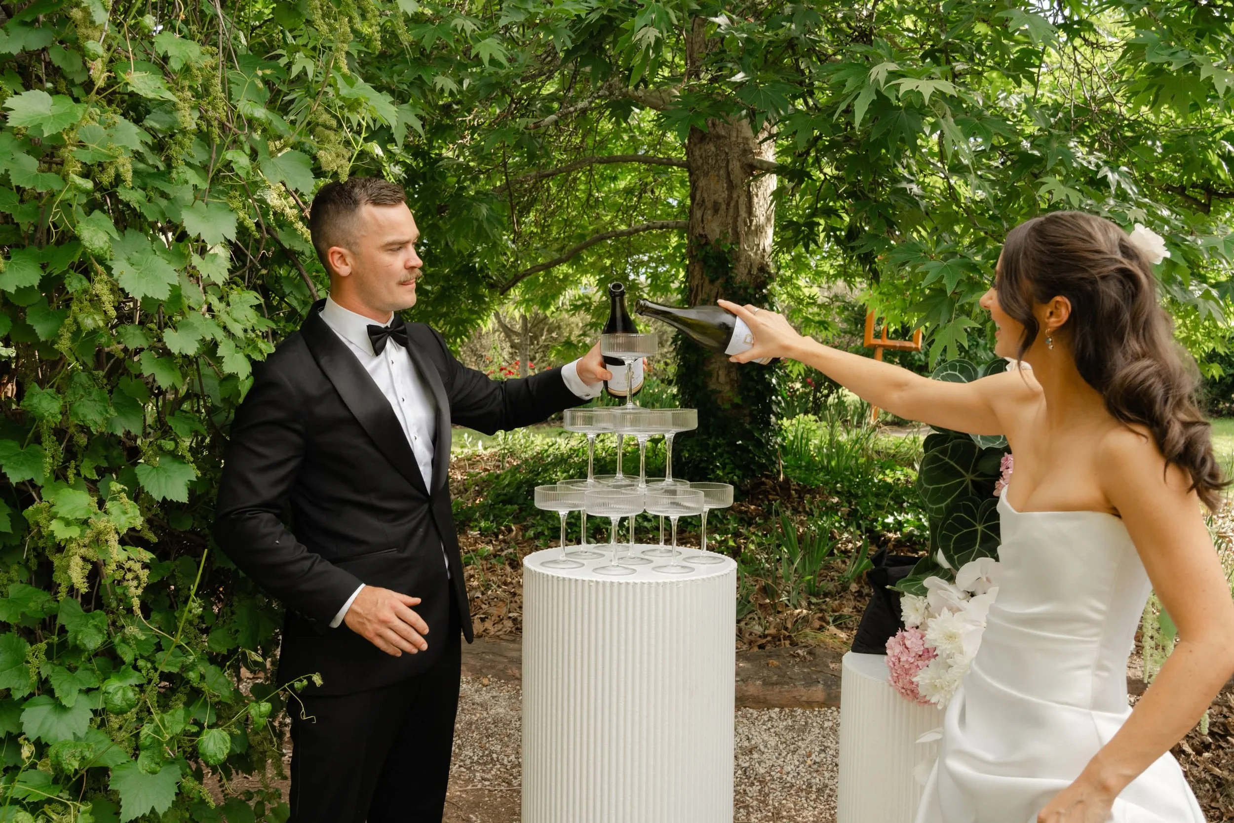 A man in a black tuxedo with a bowtie toasting with a woman in a white dress outdoors during the daytime. The woman is reaching across a table with champagne glasses arranged in a pyramid, under trees with green leaves.