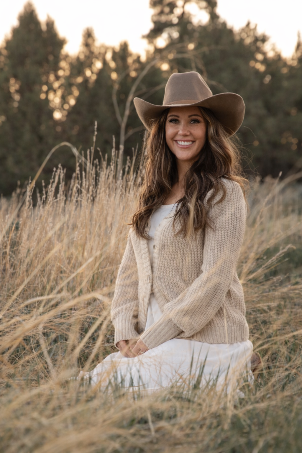 A woman with long curly brown hair and a tan cowboy hat, sitting in a field of tall dry grass near trees, smiling at the camera, wearing a beige cardigan and a white dress.