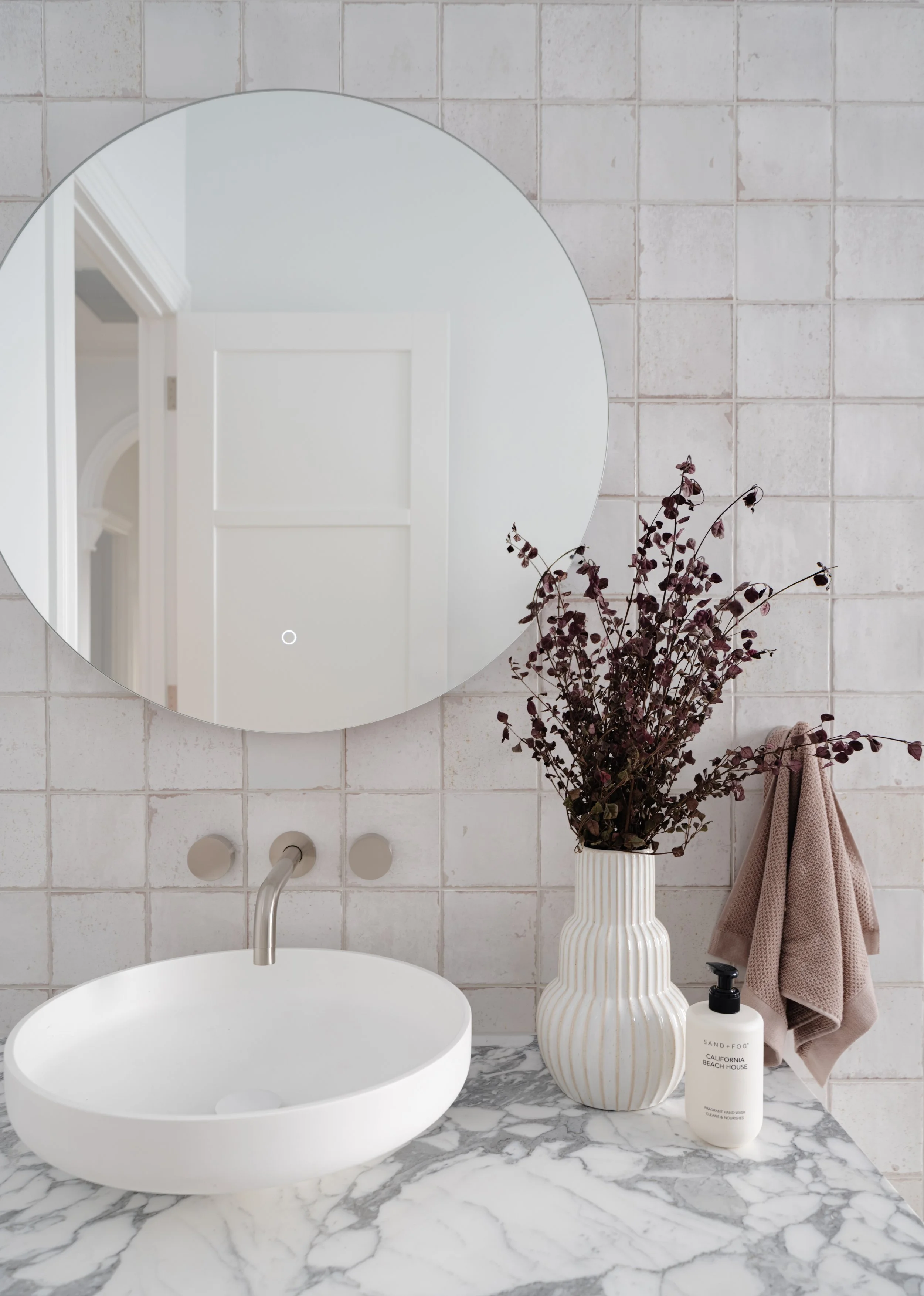 Bathroom vanity with a round mirror, white vessel sink, marble countertop, a striped vase with dark dried flowers, a beige hand towel, and a bottle of hand soap on a tiled wall background.